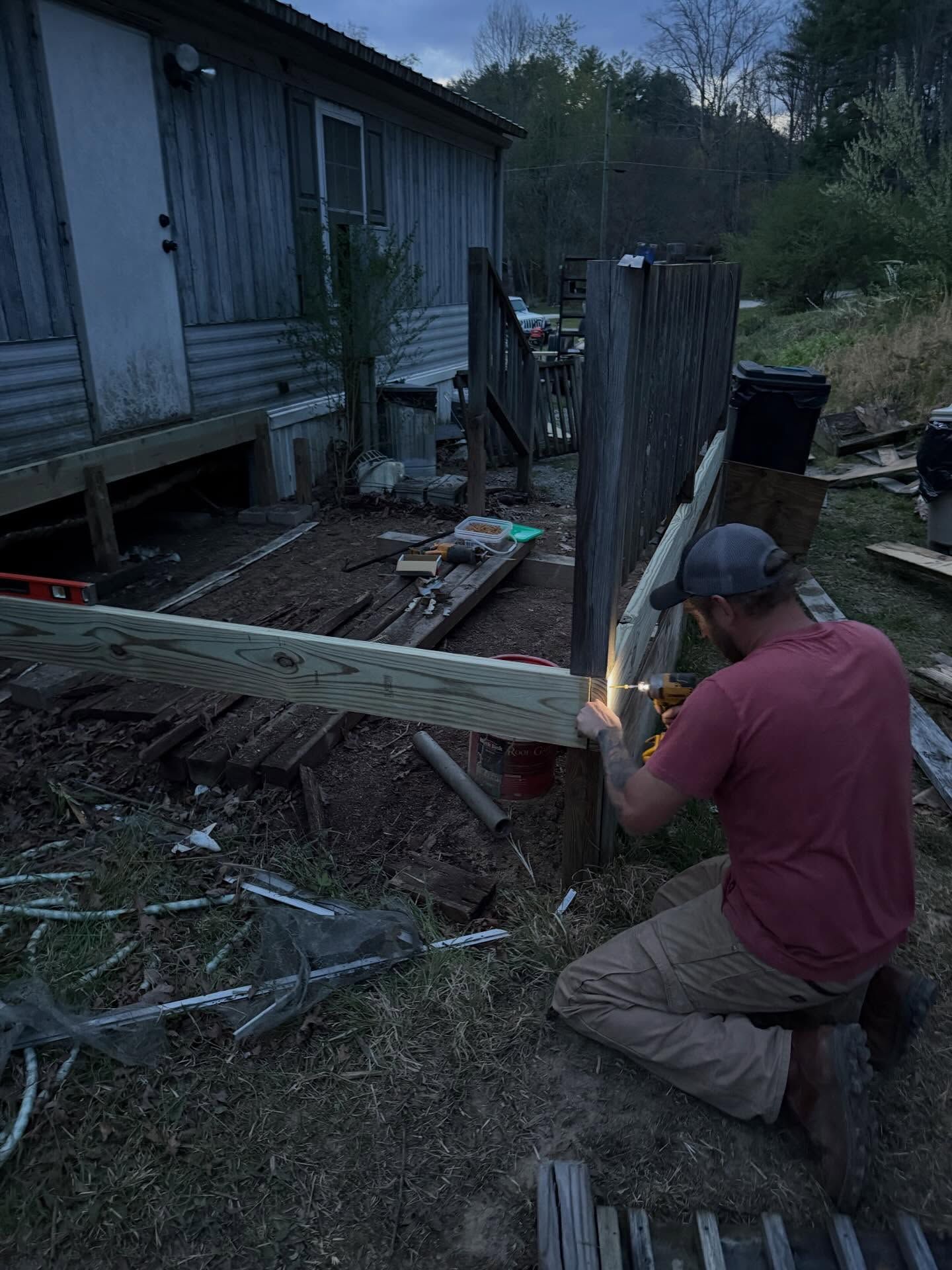 Person kneeling outdoors, cutting a wooden plank beside a weathered house at dusk.