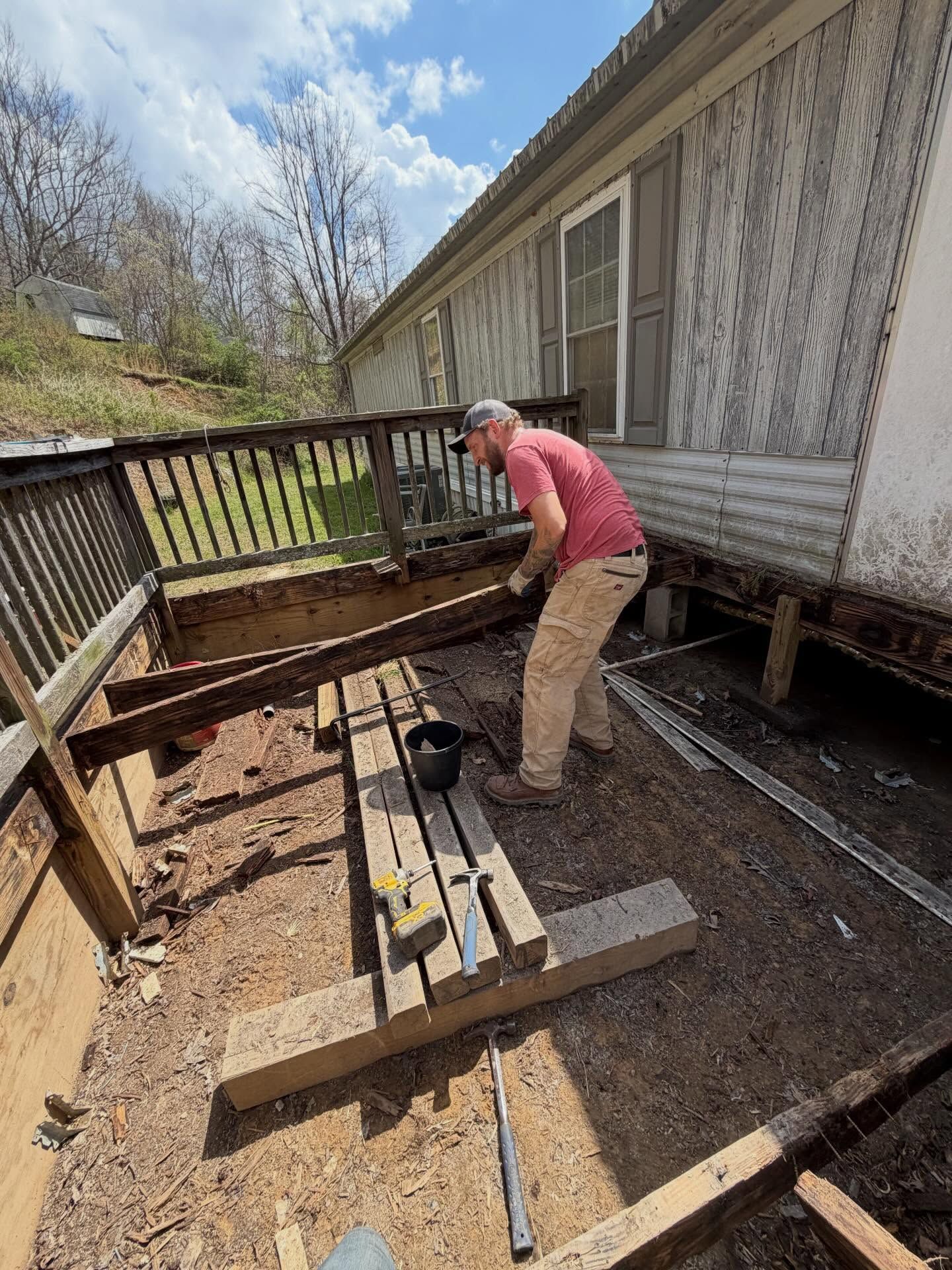 Person working on a wooden deck beside a house, surrounded by tools and lumber.