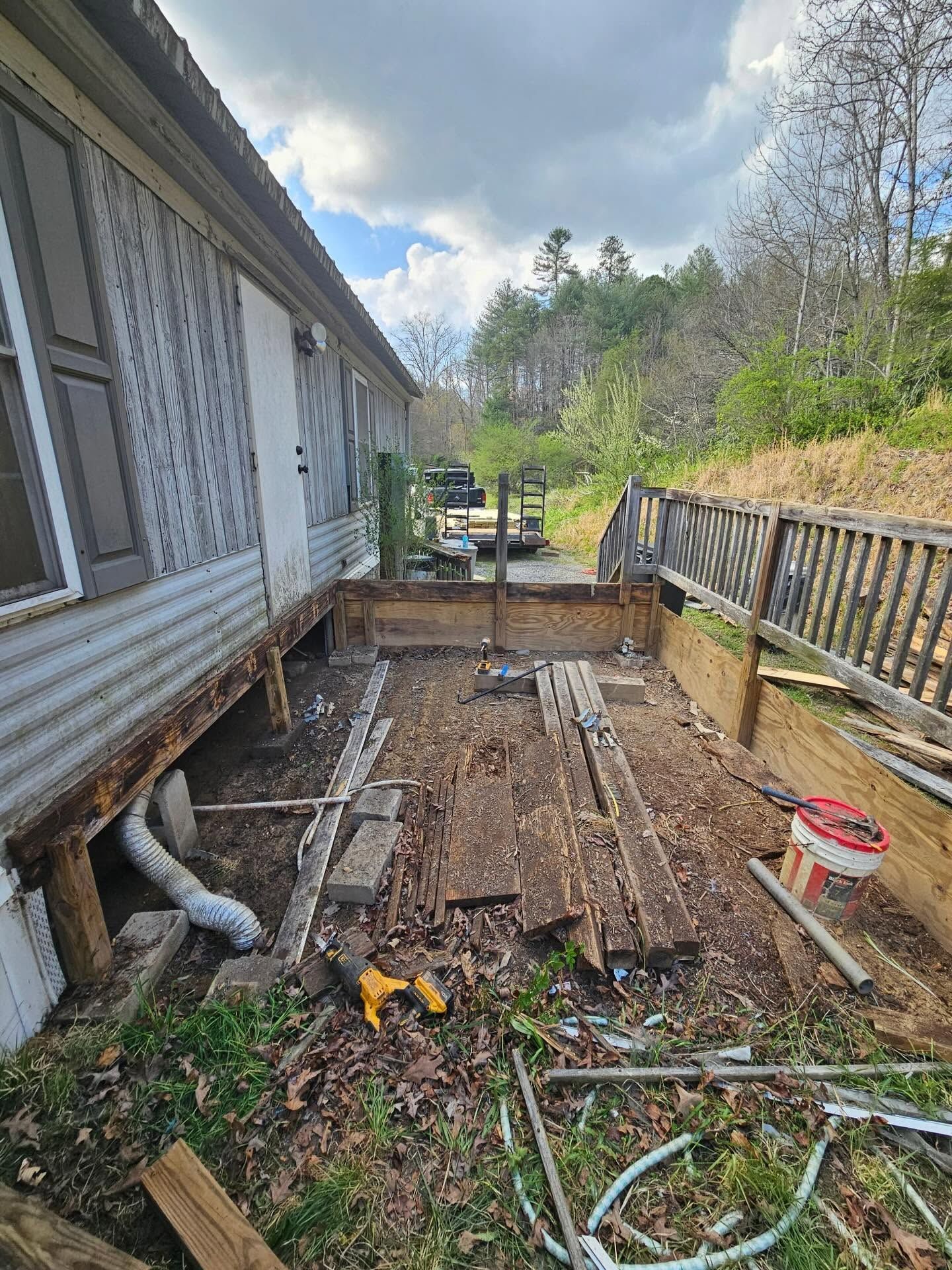 Damaged wooden deck beside a house, with scattered debris and railing along a wooded slope.