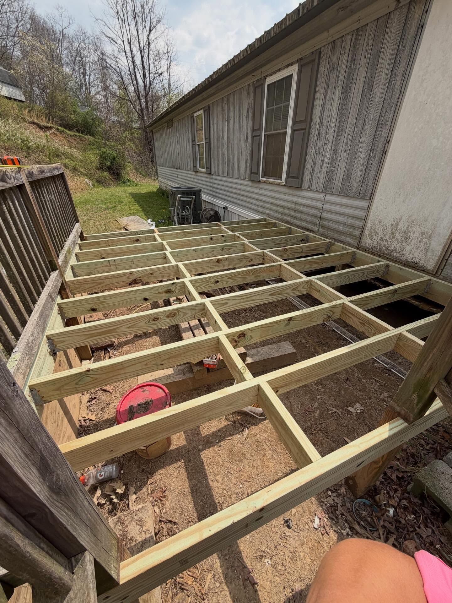 Raised deck frame under construction beside a house, with exposed joists and dirt ground below.