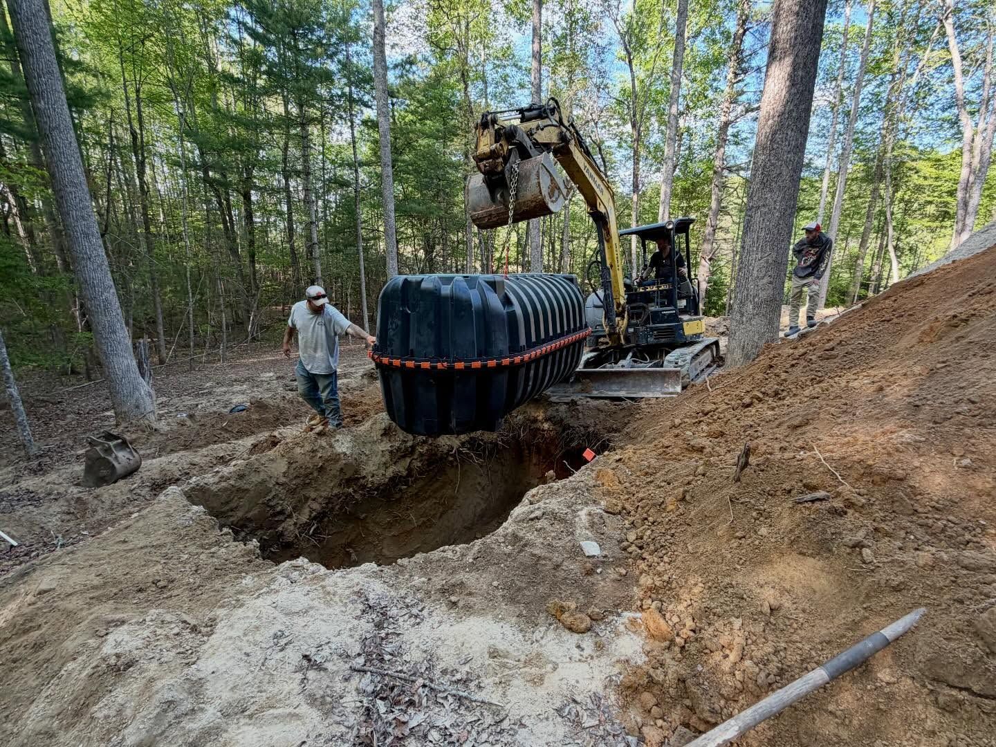 Construction equipment lifting a large black tank into a deep trench in a wooded area