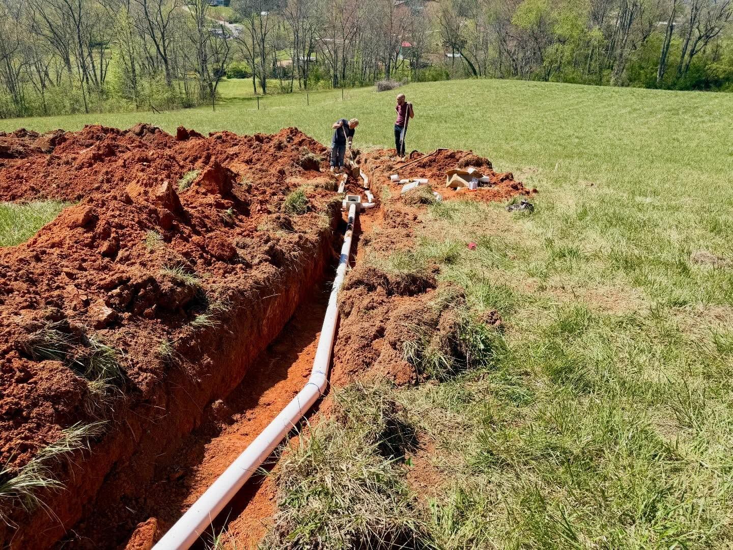 Trench with white pipe laid in red soil beside a grassy field, with two workers in the background