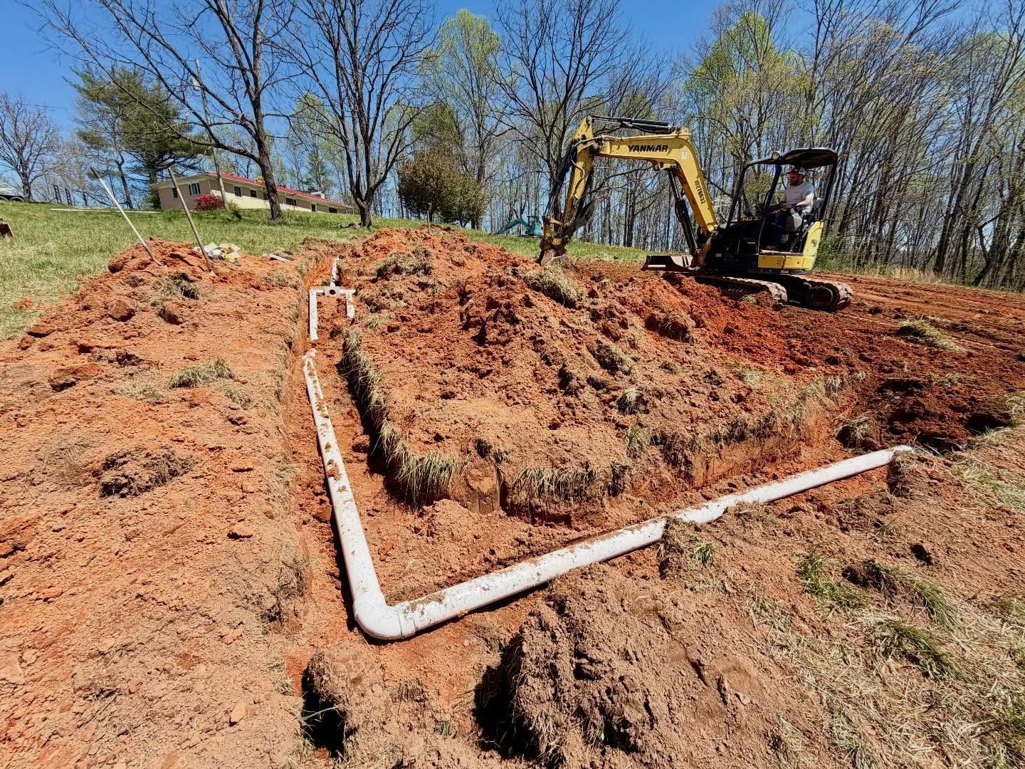 Excavator digging a trench beside white PVC pipes on a dirt hillside under a blue sky