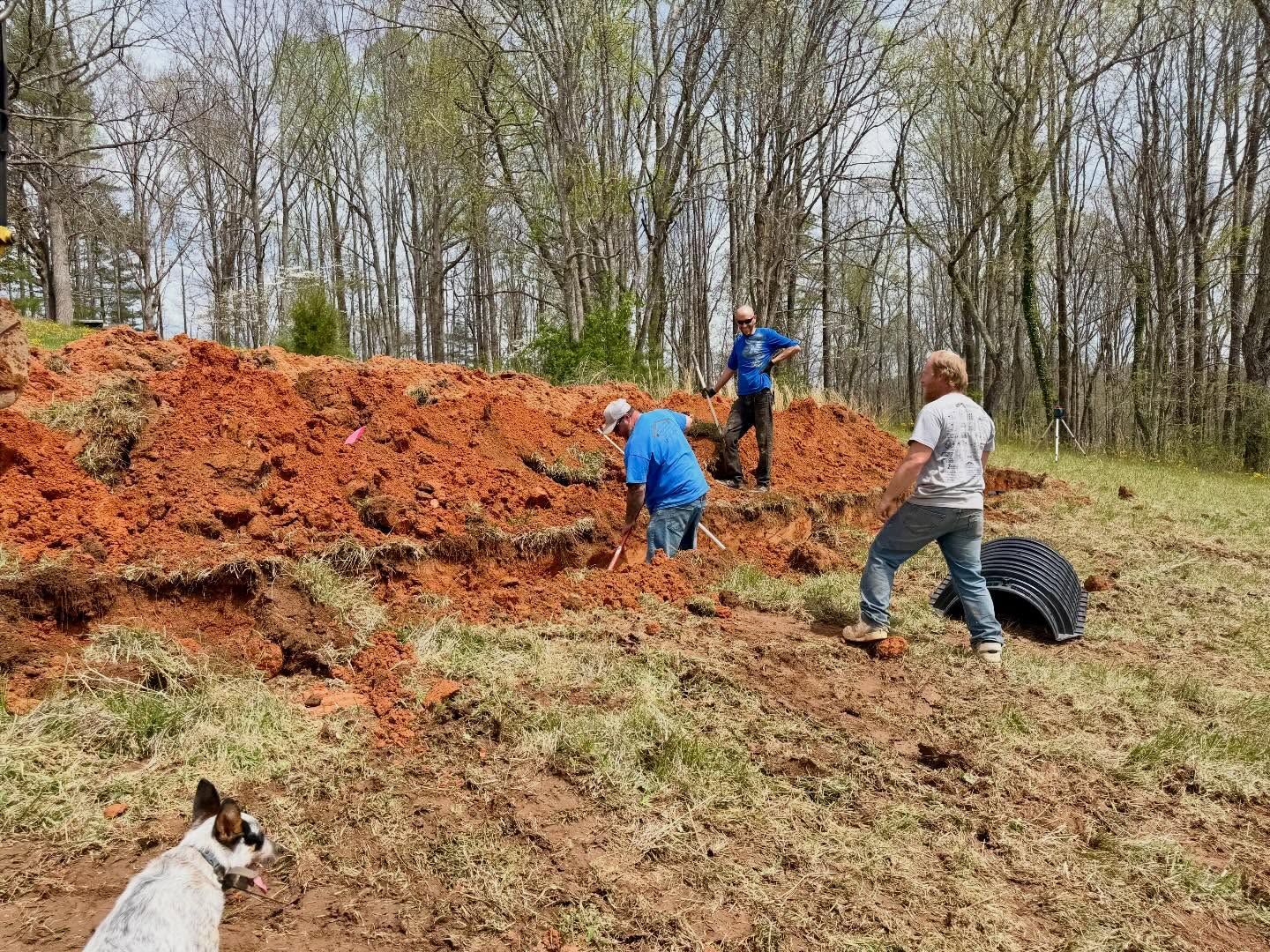 Three people working on a red dirt slope beside a wooded area, with a dog in the foreground.