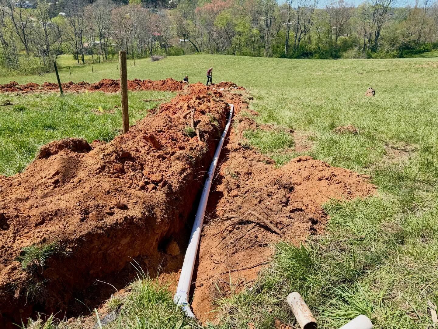 Trenched ditch with exposed white pipe in a grassy field, with piles of reddish soil nearby