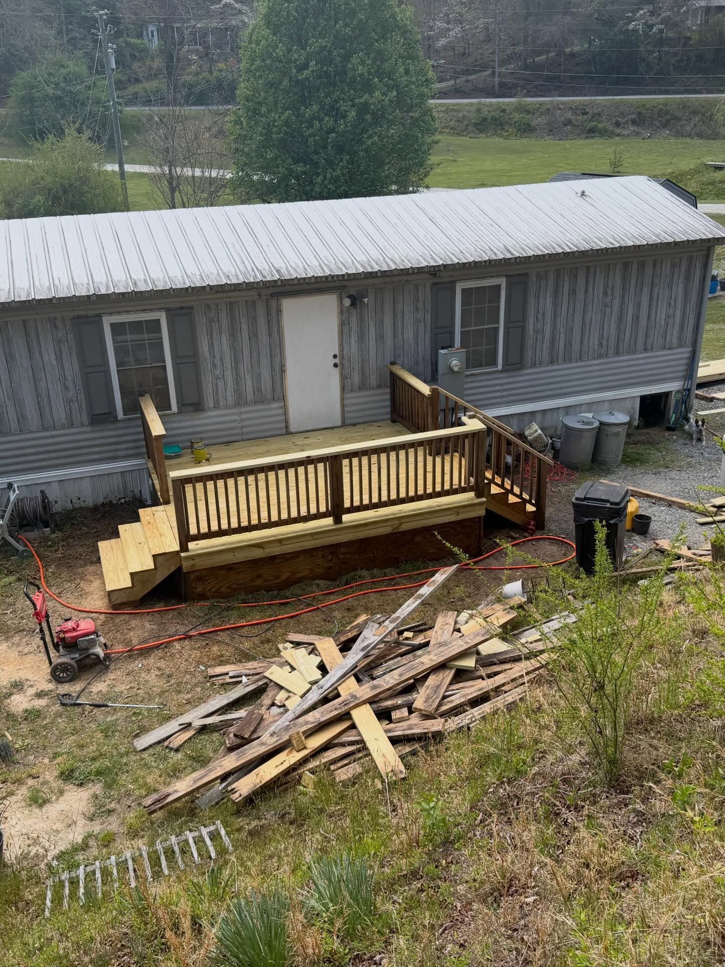 Mobile home with a new wooden deck and stairs, surrounded by scattered construction lumber in a yard.