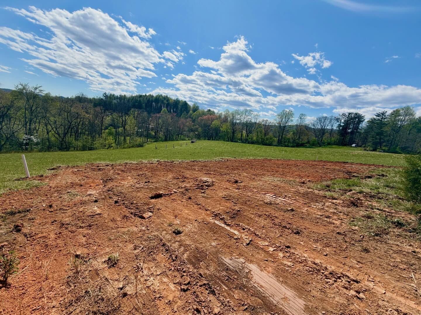 Bare dirt clearing in a grassy field under a blue sky with clouds, bordered by trees