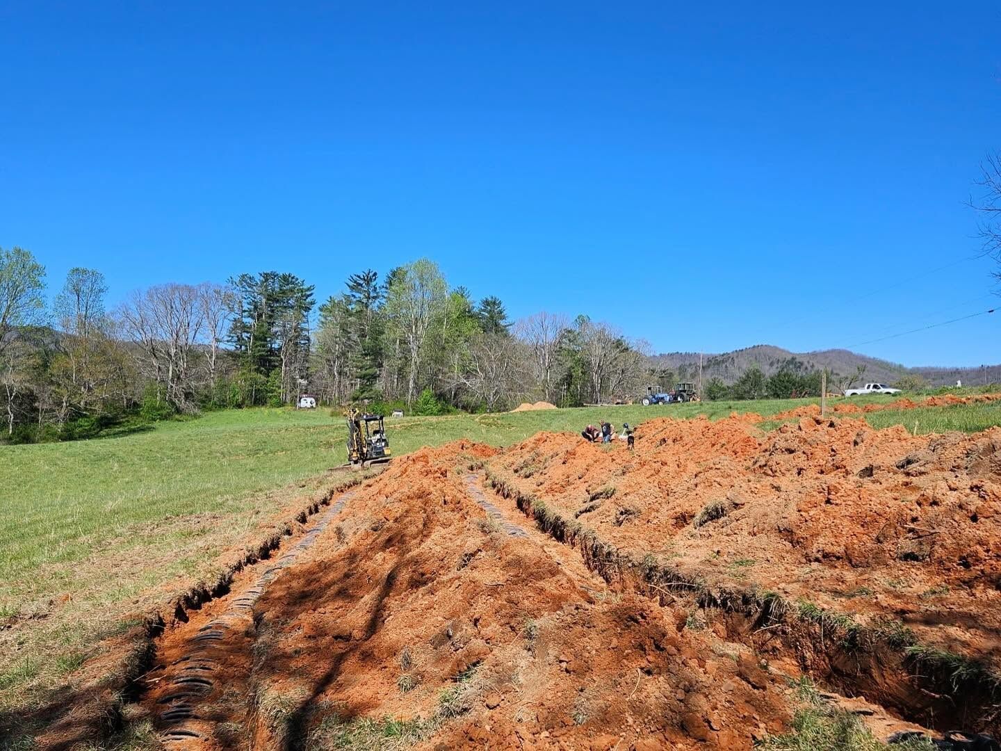 Dirt field with tire tracks beside a grassy meadow under a clear blue sky, with trees and hills in the distance