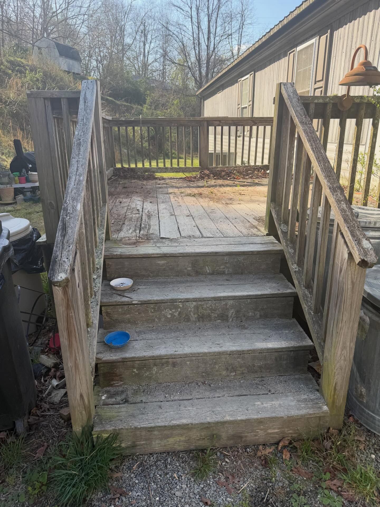 Weathered wooden steps and railing leading to a small deck beside a house, with trees in the background.