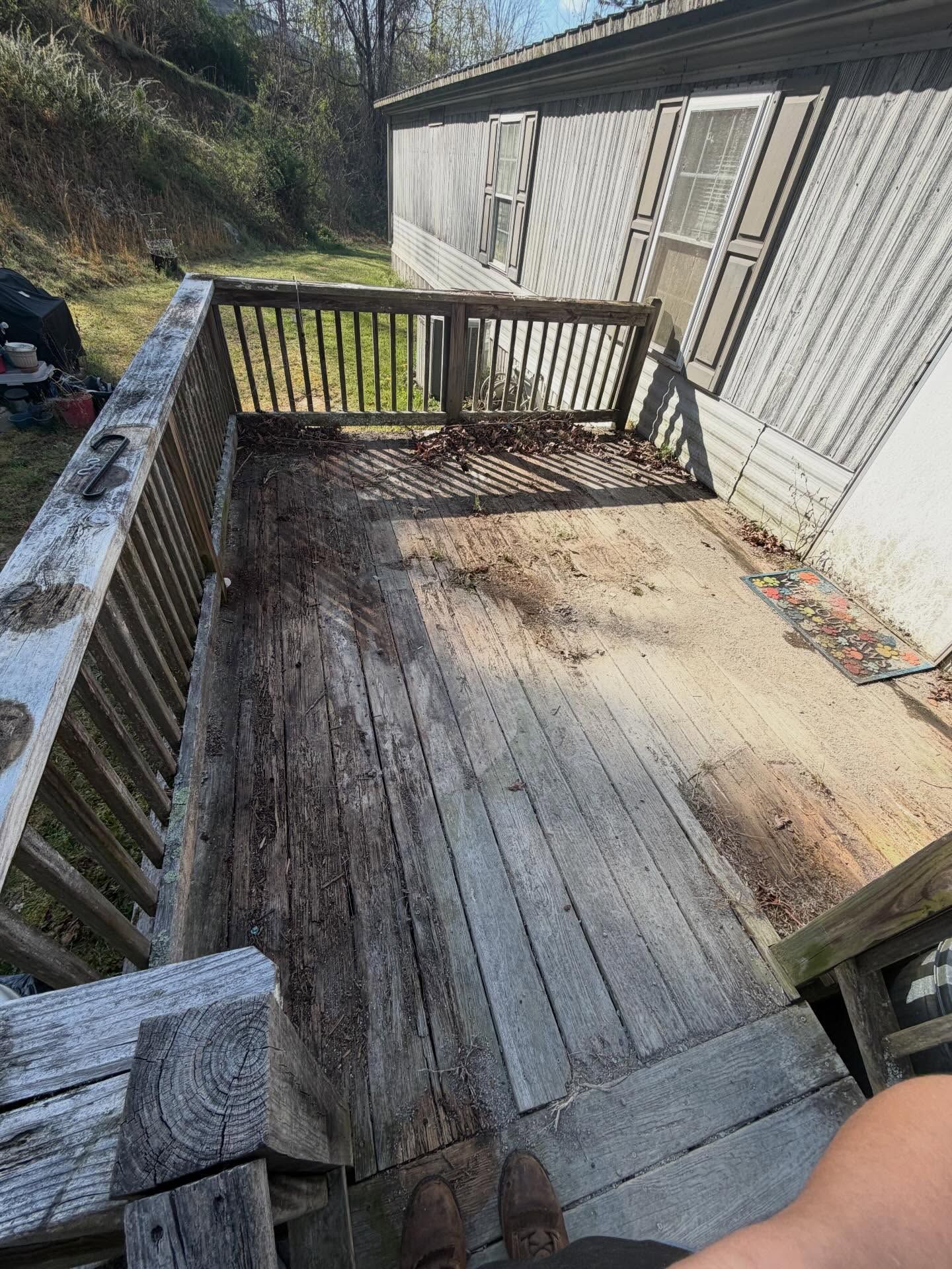 Wooden deck and railing beside a white building, with weathered boards and a wooded yard beyond.