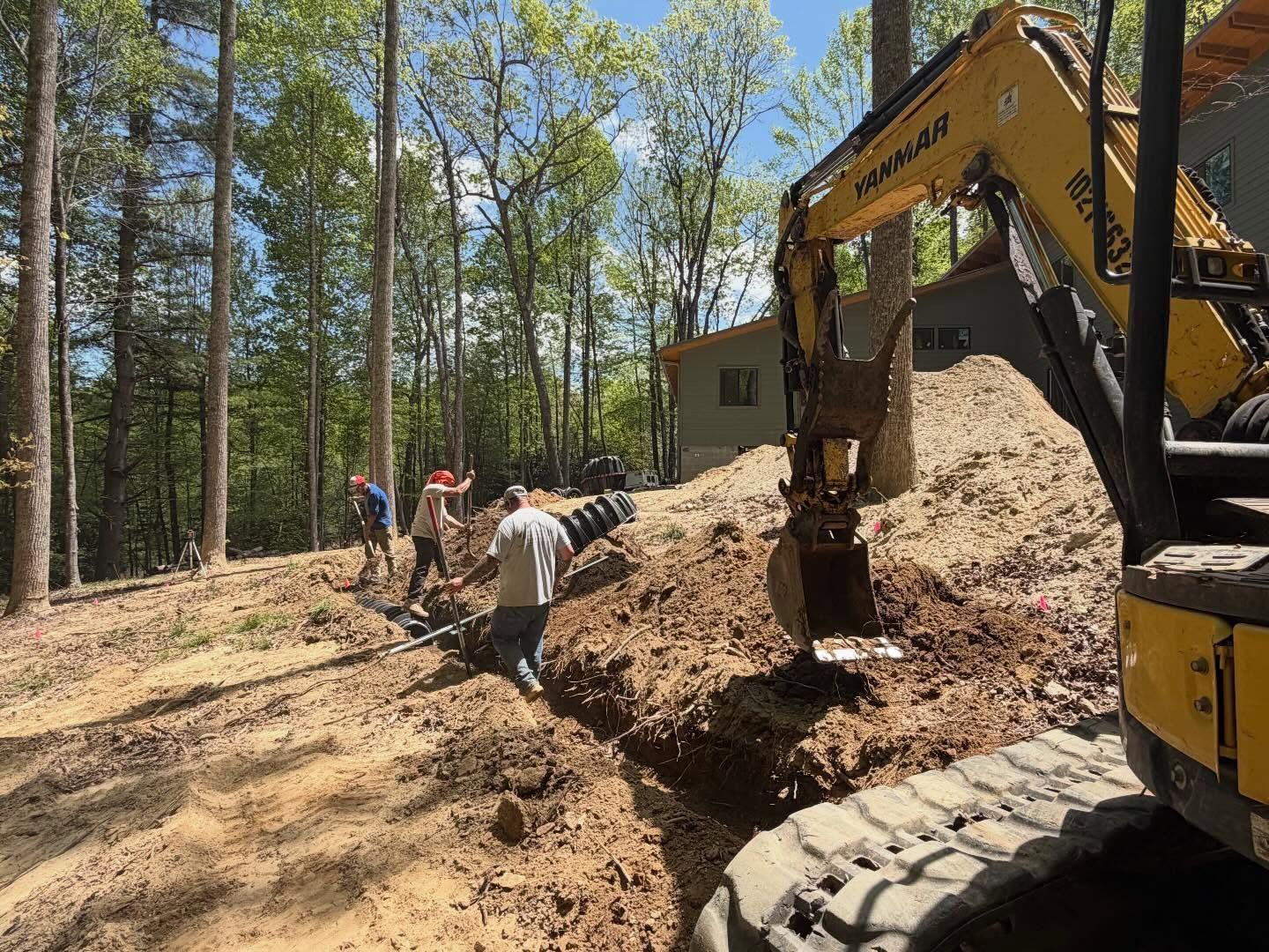 Construction workers and an excavator moving a large dirt pile in a wooded area