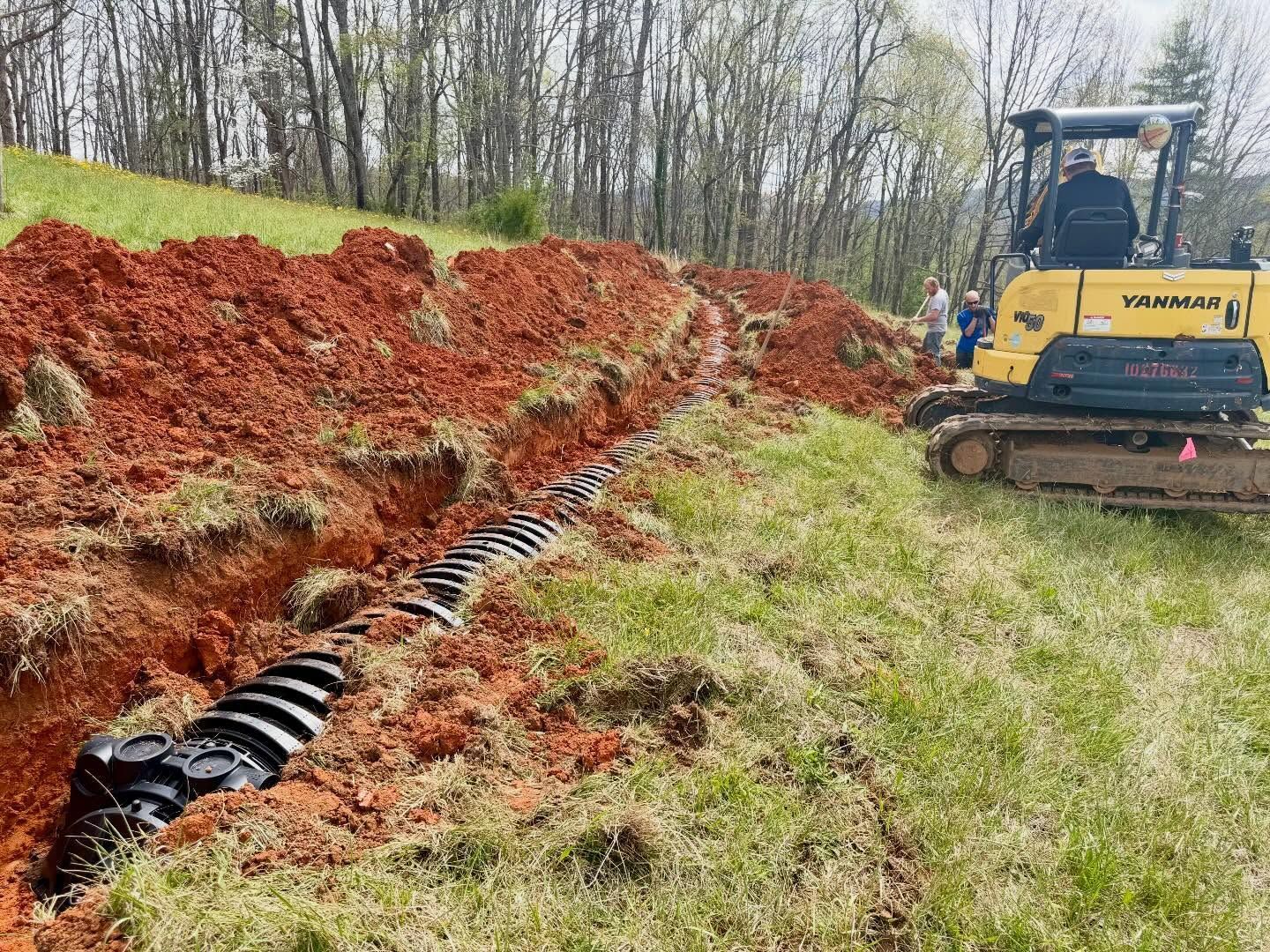 Yellow excavator beside red soil trench with black drainage pipe on a grassy slope under bare trees