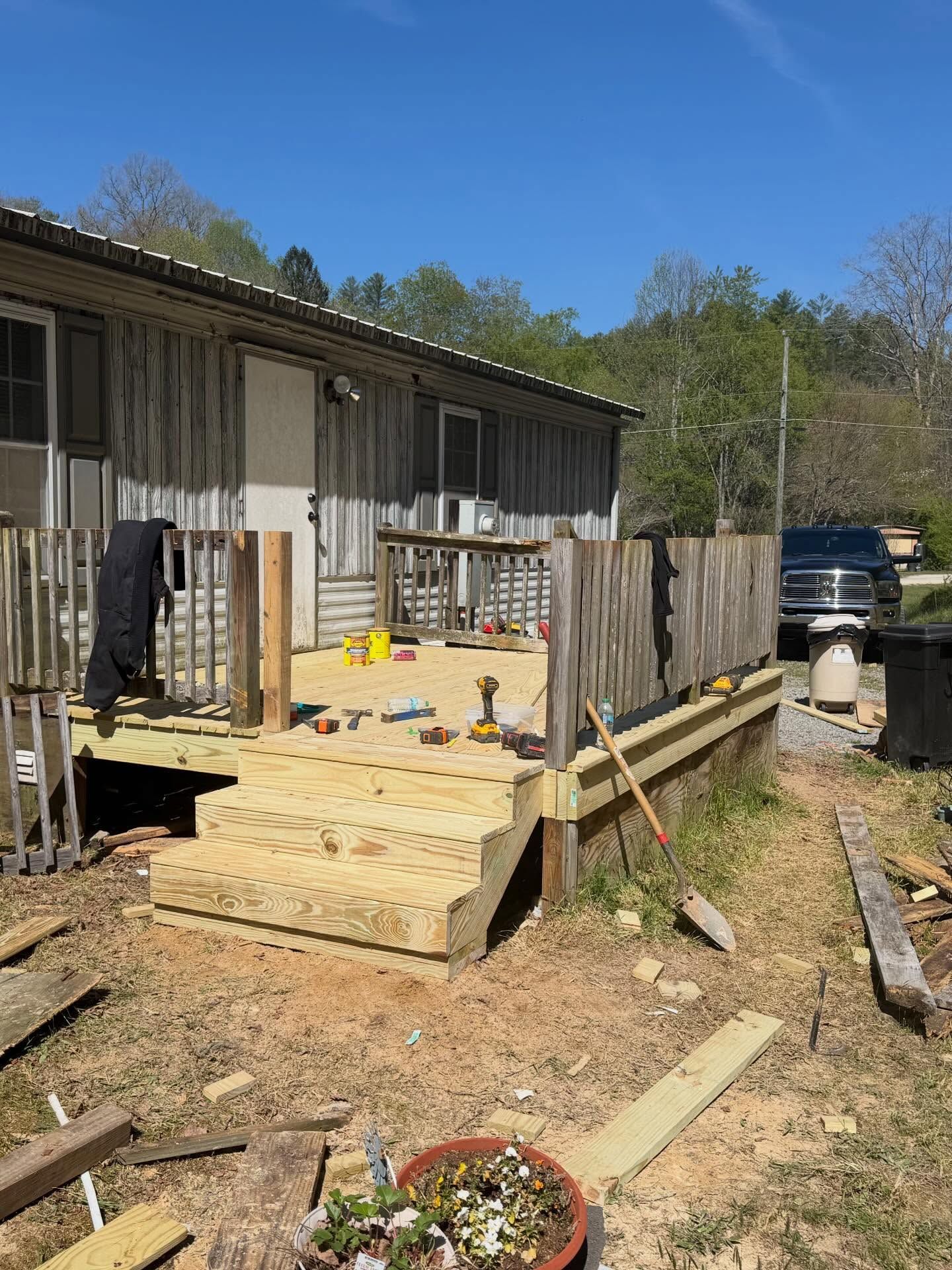 Wooden deck construction beside a house, with tools, fencing, and lumber in a sunny yard.