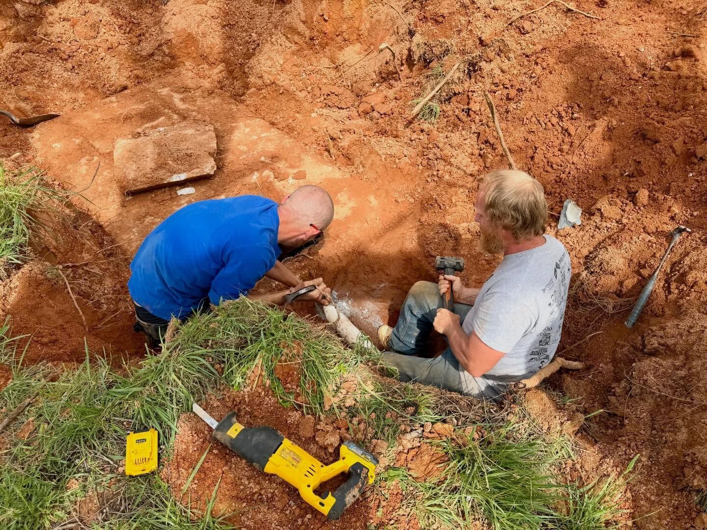 Two workers crouch in a dirt excavation next to a yellow power tool and shovel.