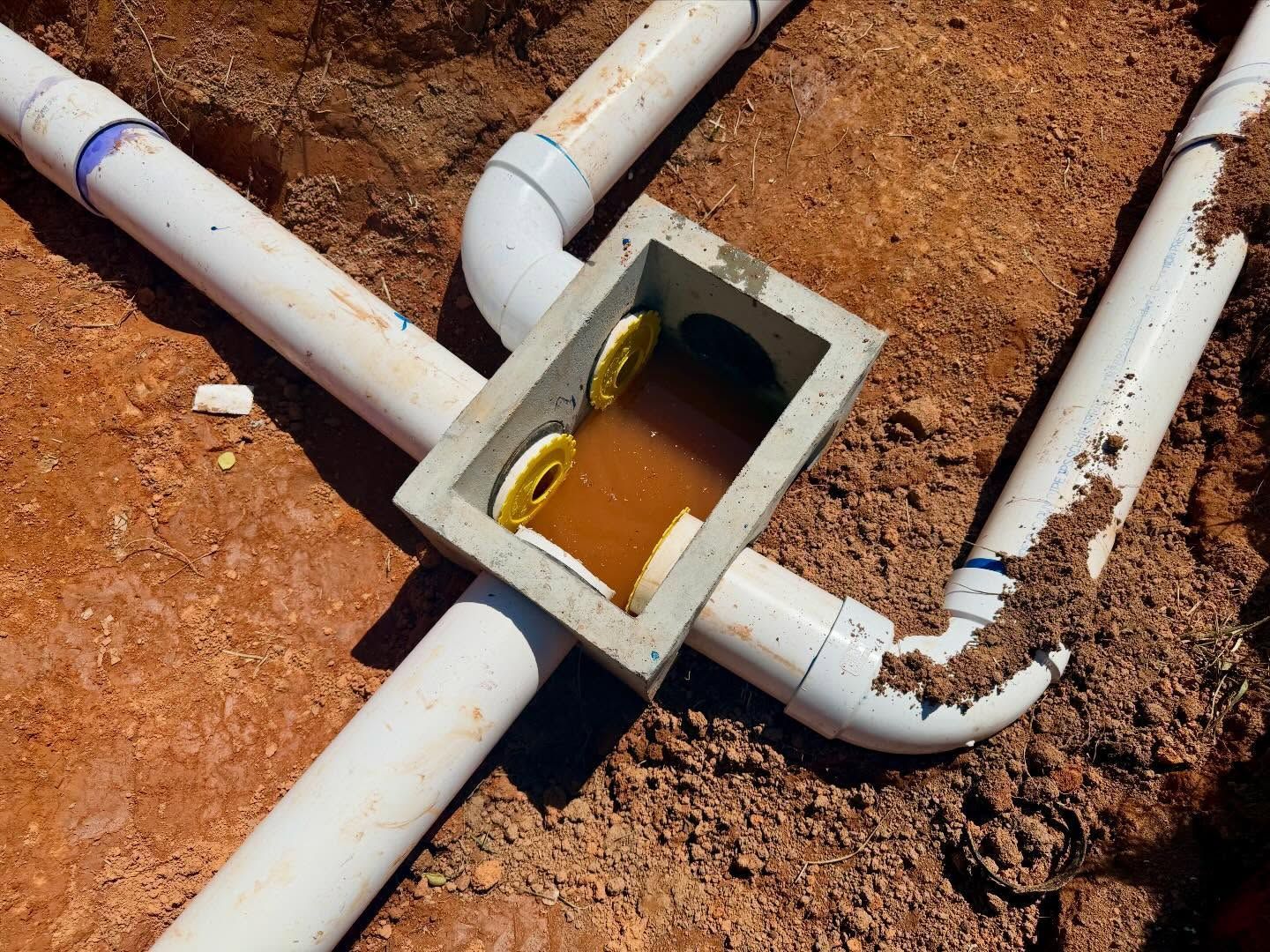 PVC pipes leading to a small concrete inspection box in reddish dirt