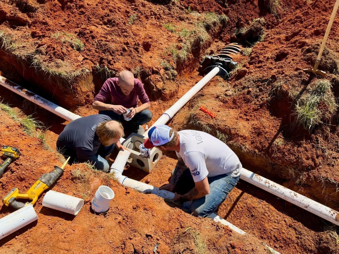 Three workers repairing white PVC pipes in a red dirt trench outdoors