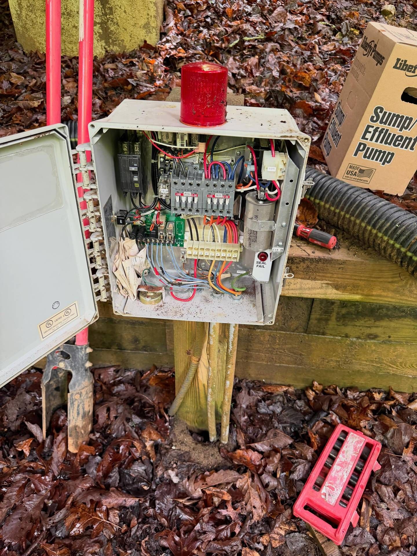 Open electrical control box with red beacon light beside a sump pump in a wet wooded area