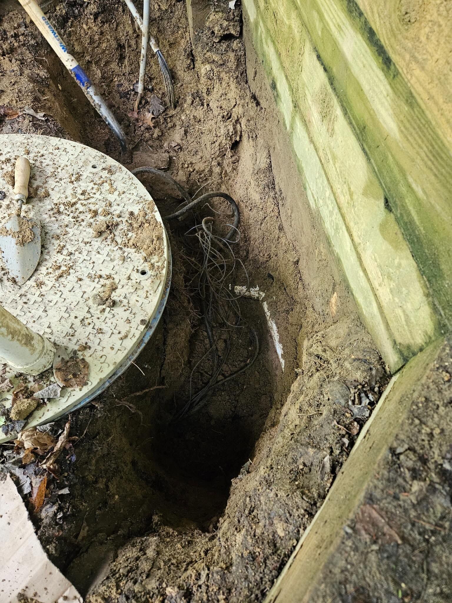 Open soil trench beside a wooden fence, with a round utility cover at left.