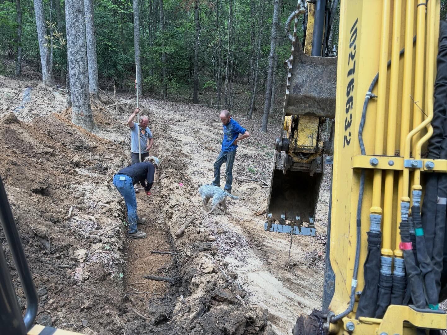 Workers digging a muddy trench beside a yellow excavator in a wooded area