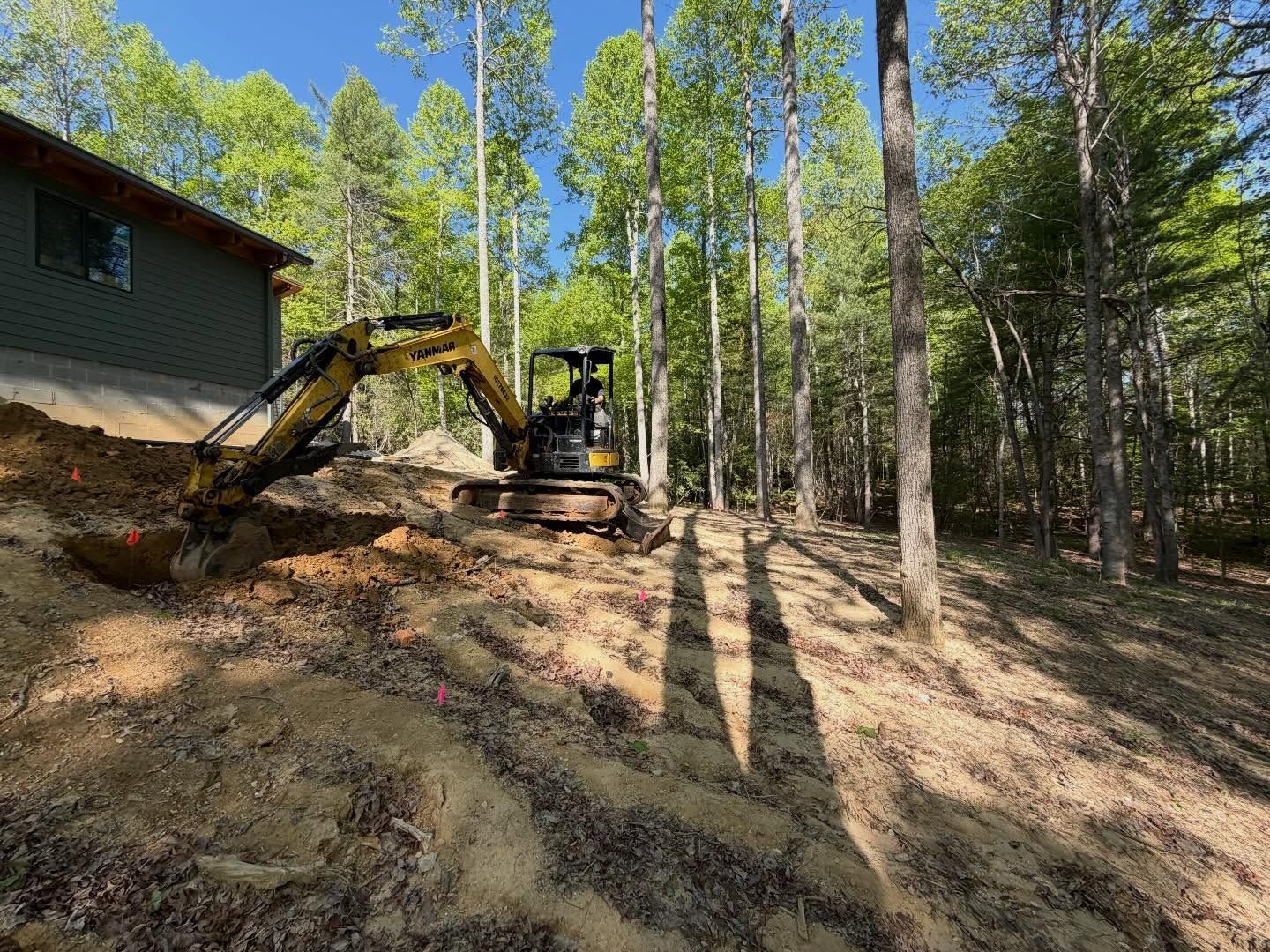 Excavator grading a dirt hillside beside a house in a wooded area, with tall trees casting long shadows.