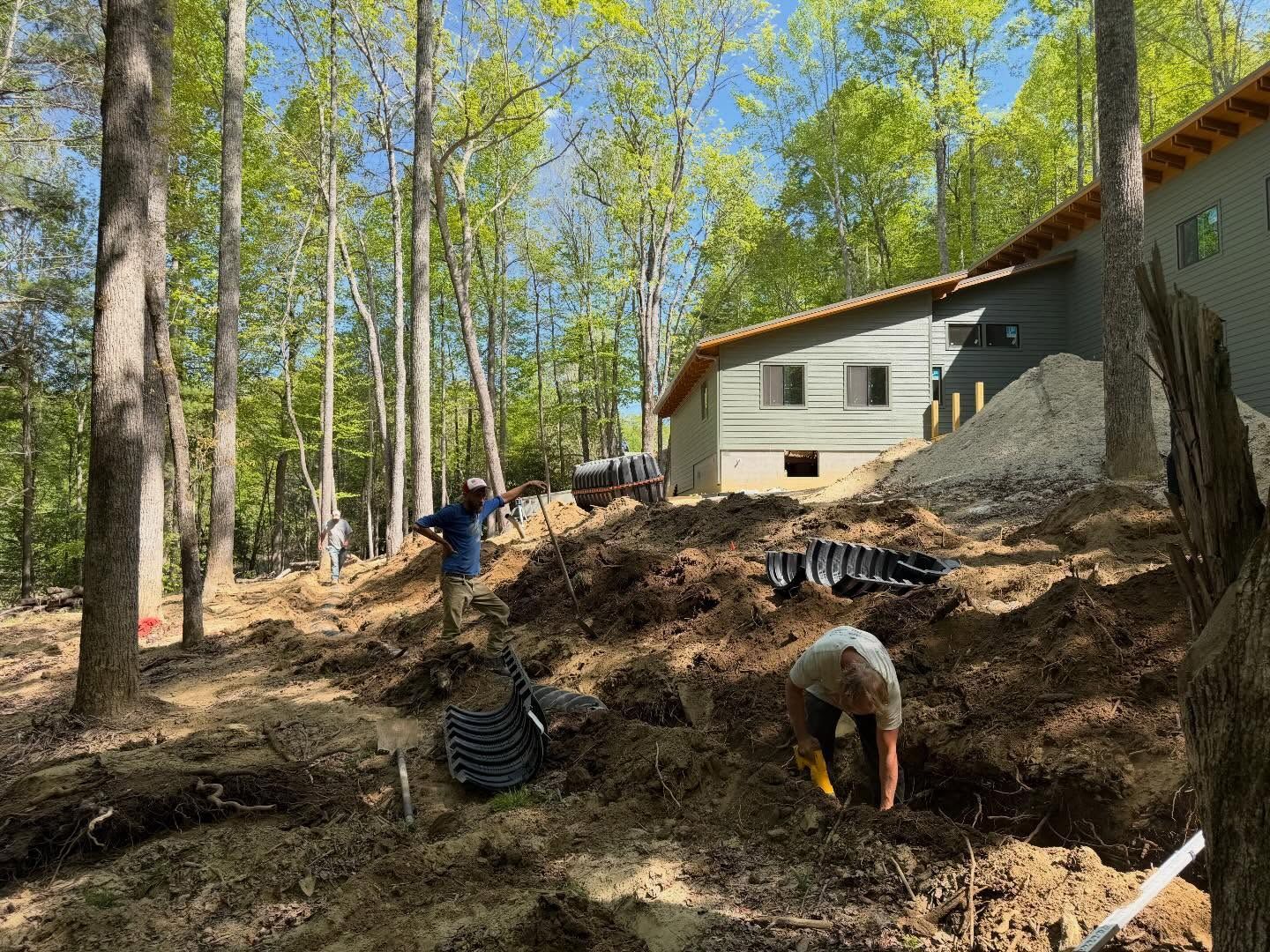 People working on a wooded hillside beside a house, with trees, dirt, and scattered materials.