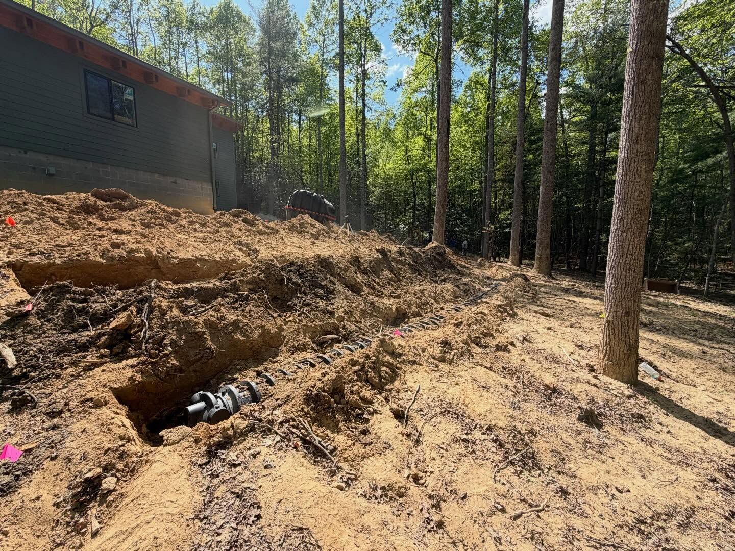 Wooded lot with a dug trench, exposed pipes, and a house beside a sandy dirt slope