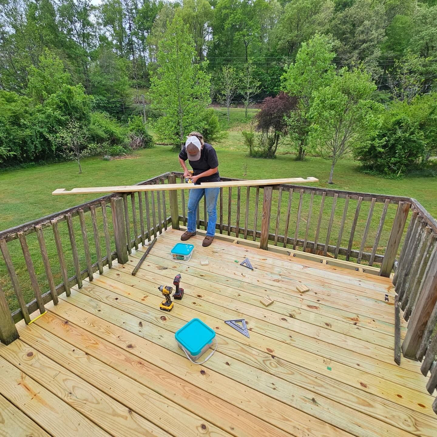 Person sanding a wooden deck outdoors with tools nearby and trees in the background