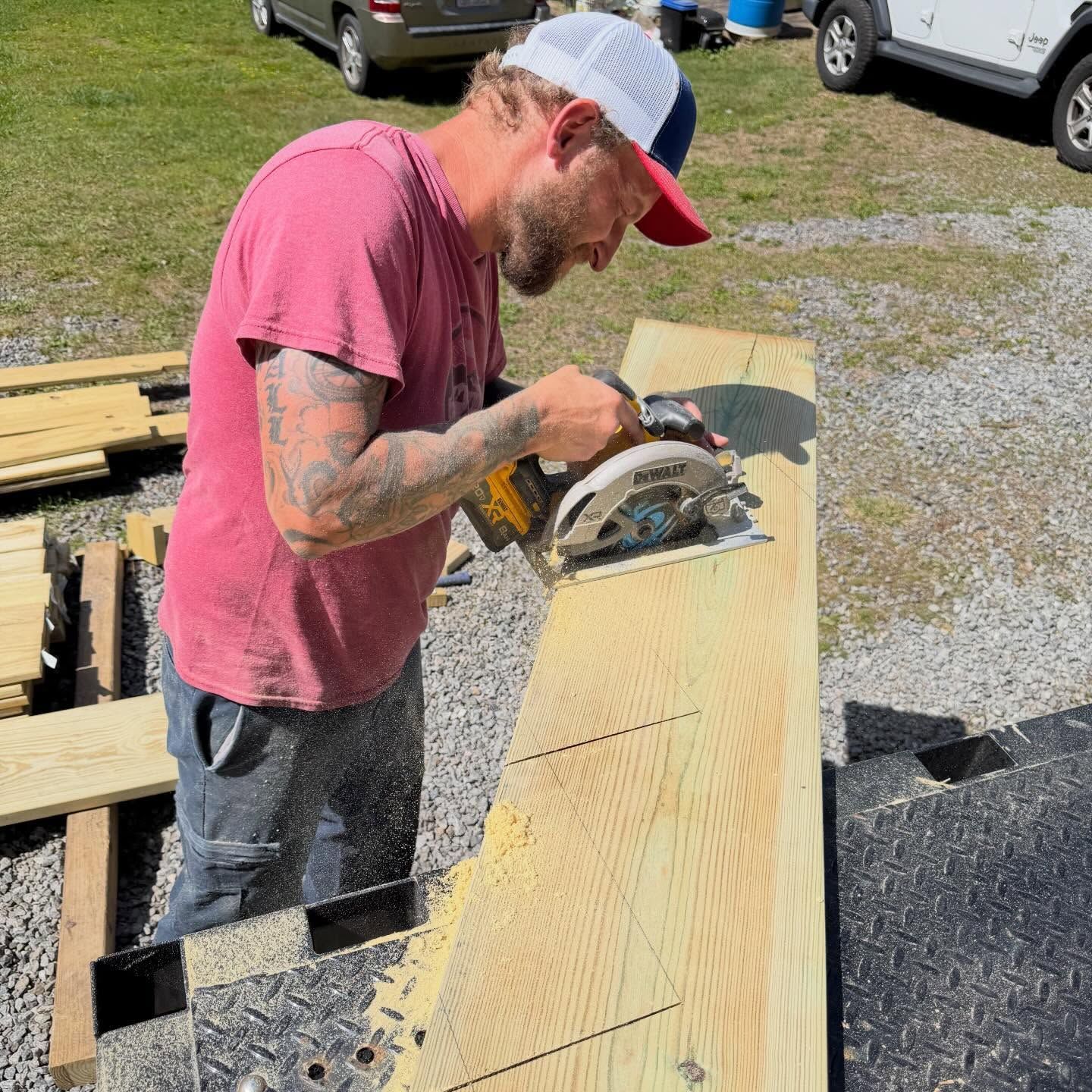 Worker in a pink shirt cutting a long wooden plank outdoors with a circular saw.