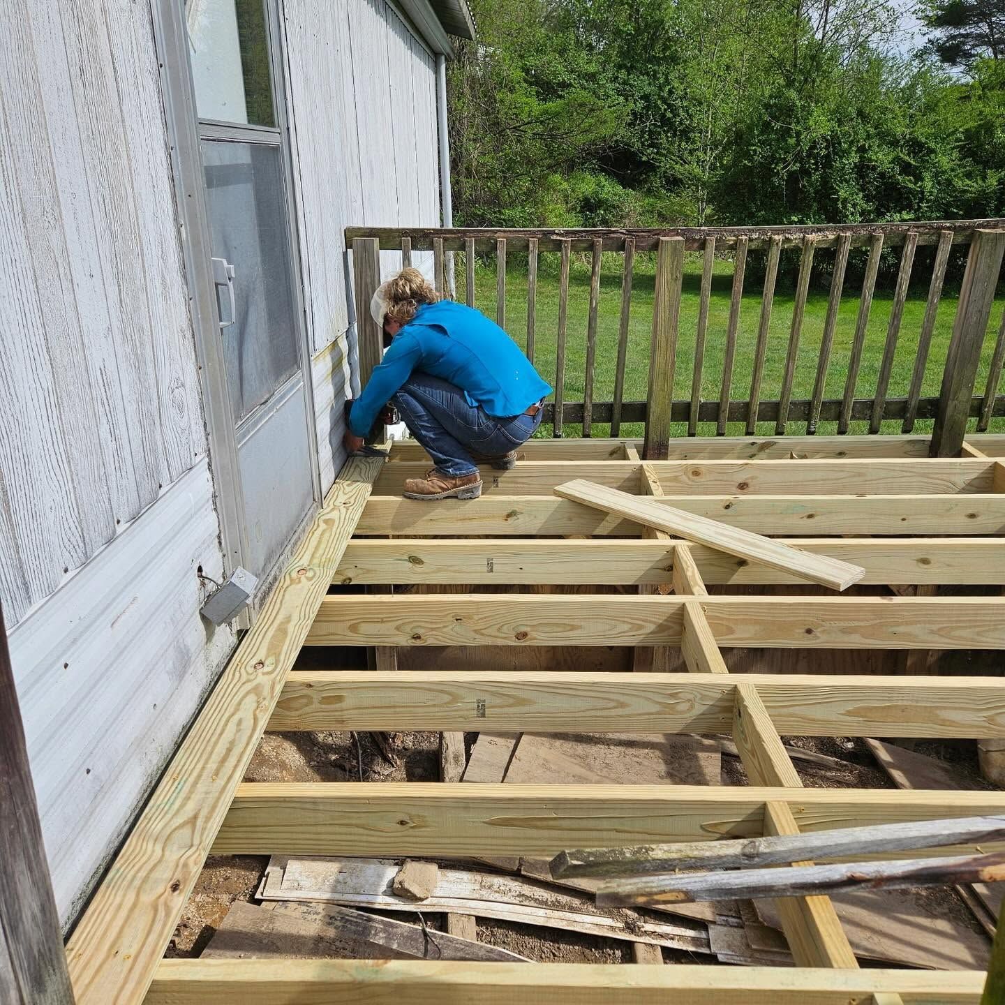 Worker crouching beside a house, repairing a wooden deck frame with exposed joists and scattered lumber.