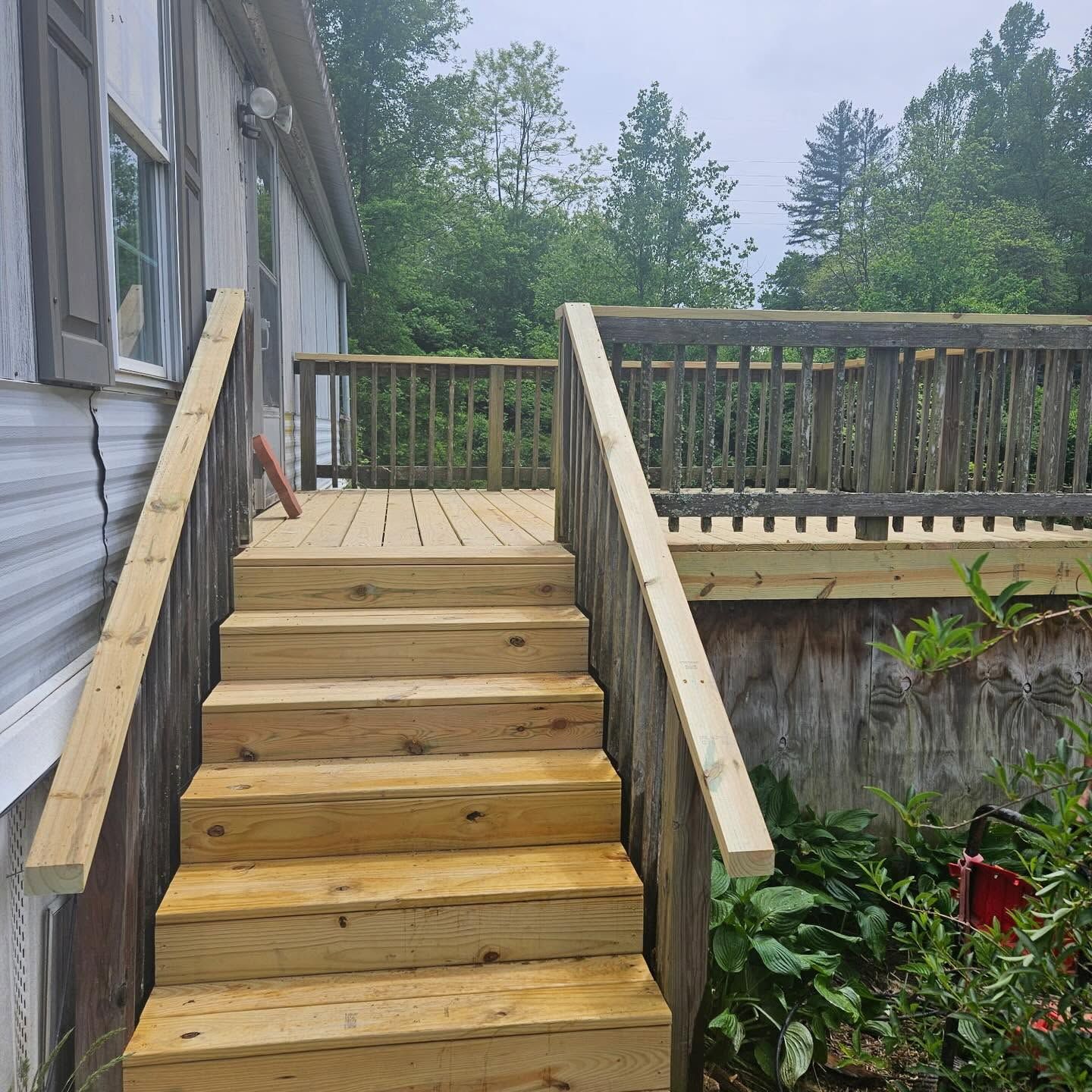 Wooden stairs leading to a raised deck beside a house, with railing and greenery around it