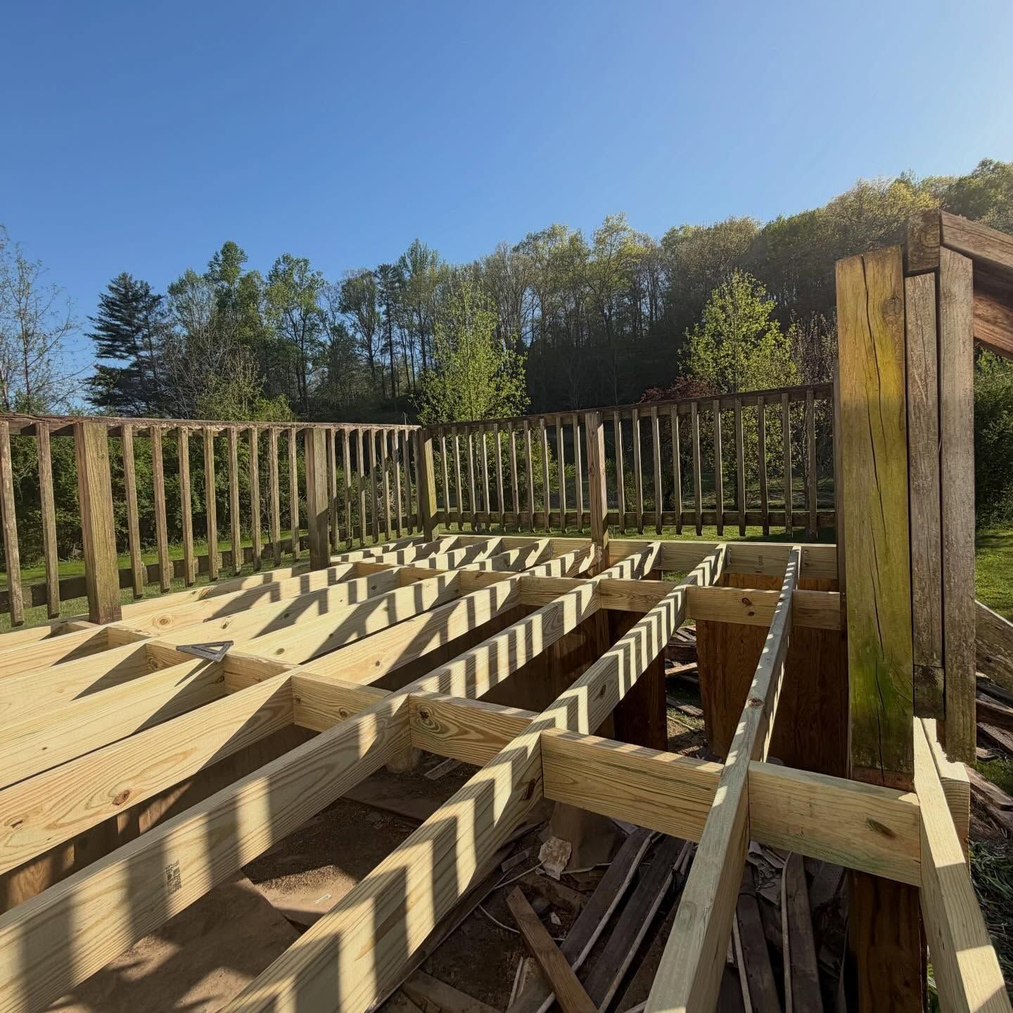 Wooden deck framing under construction with railings, set against a clear blue sky and trees.