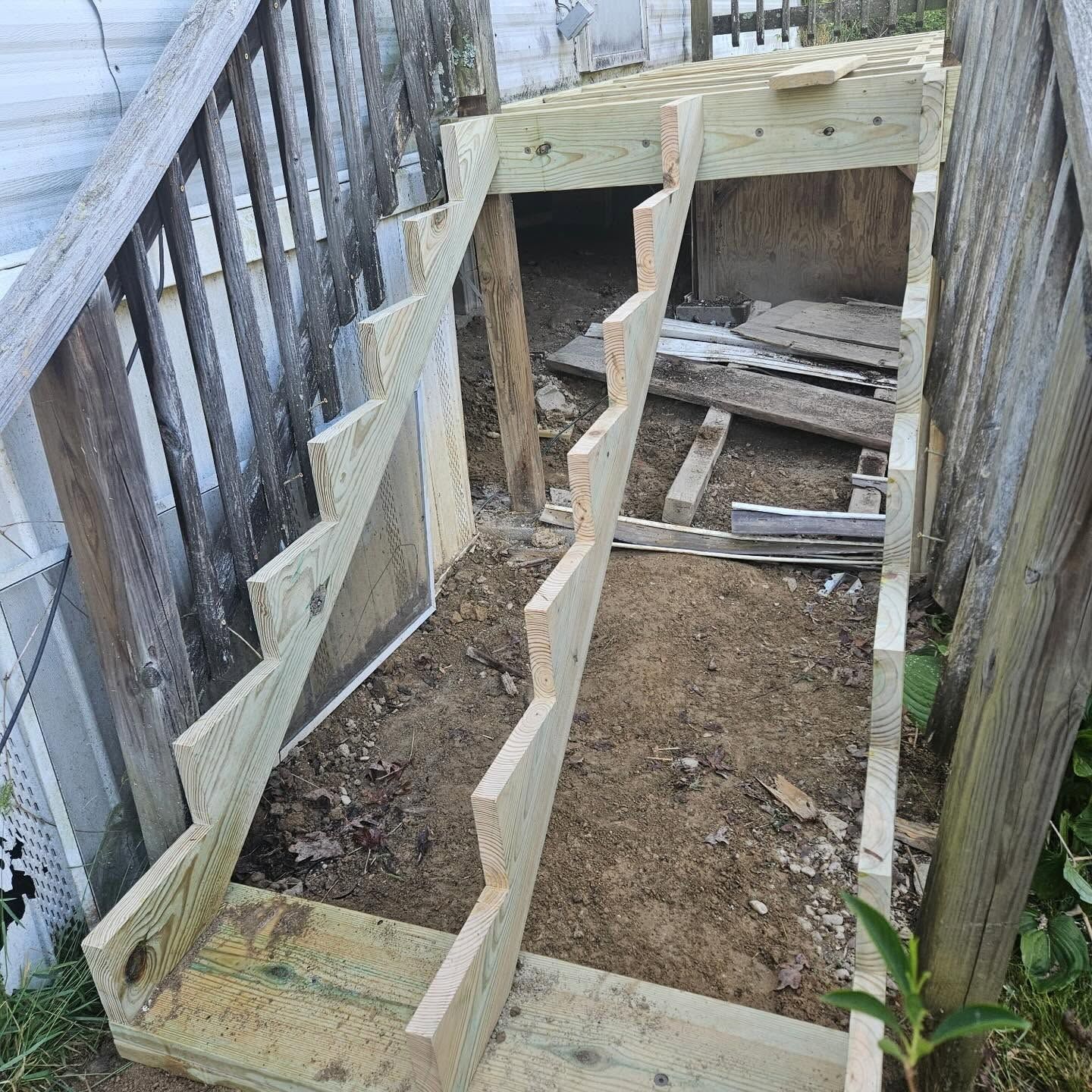 Wooden stair-frame under construction in a narrow fenced yard, with dirt ground and unfinished steps.
