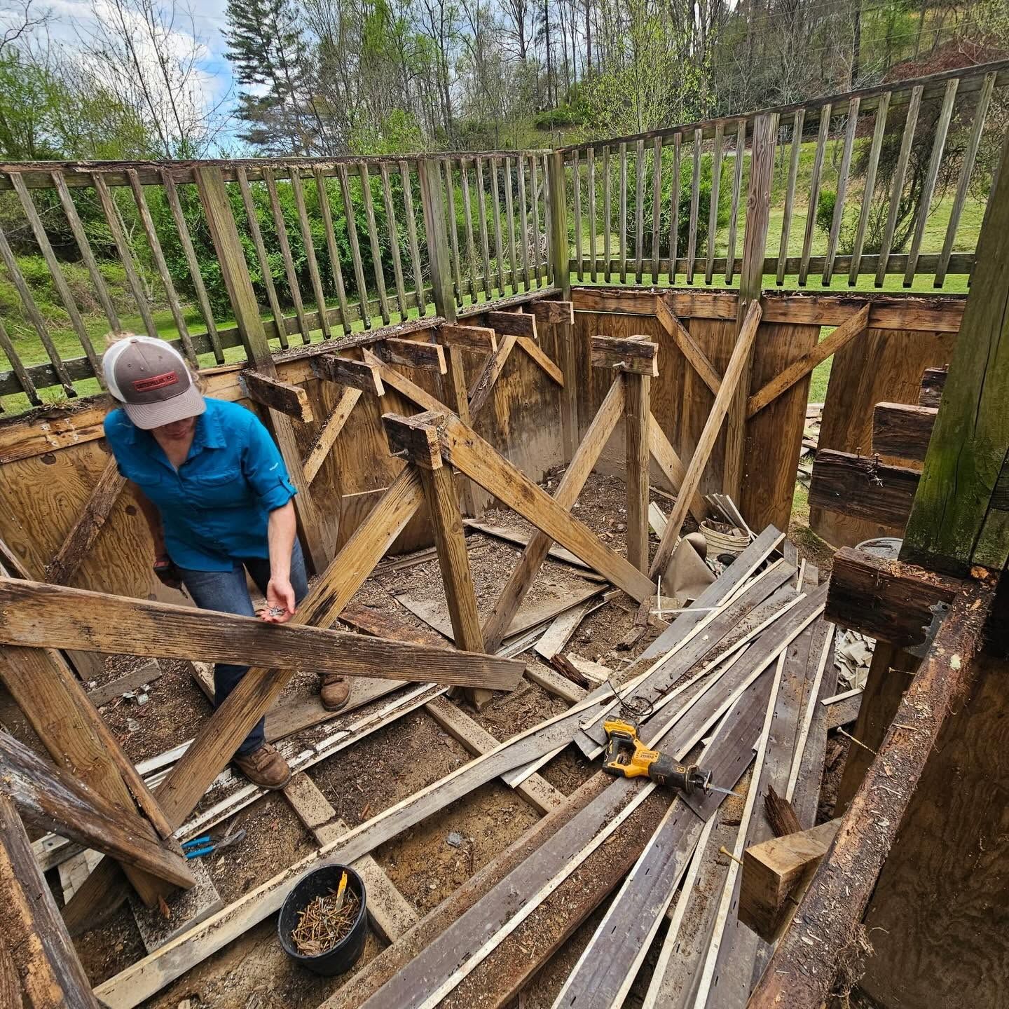 Person working in a wooden deck or patio excavation, with exposed supports and a railing above.