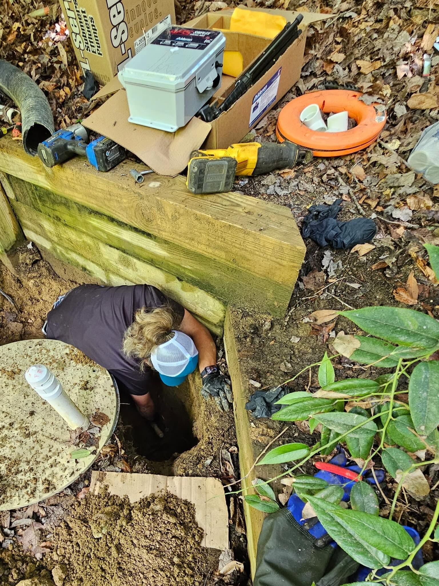 Person in a blue mask entering a small underground opening beside tools and leaves outdoors