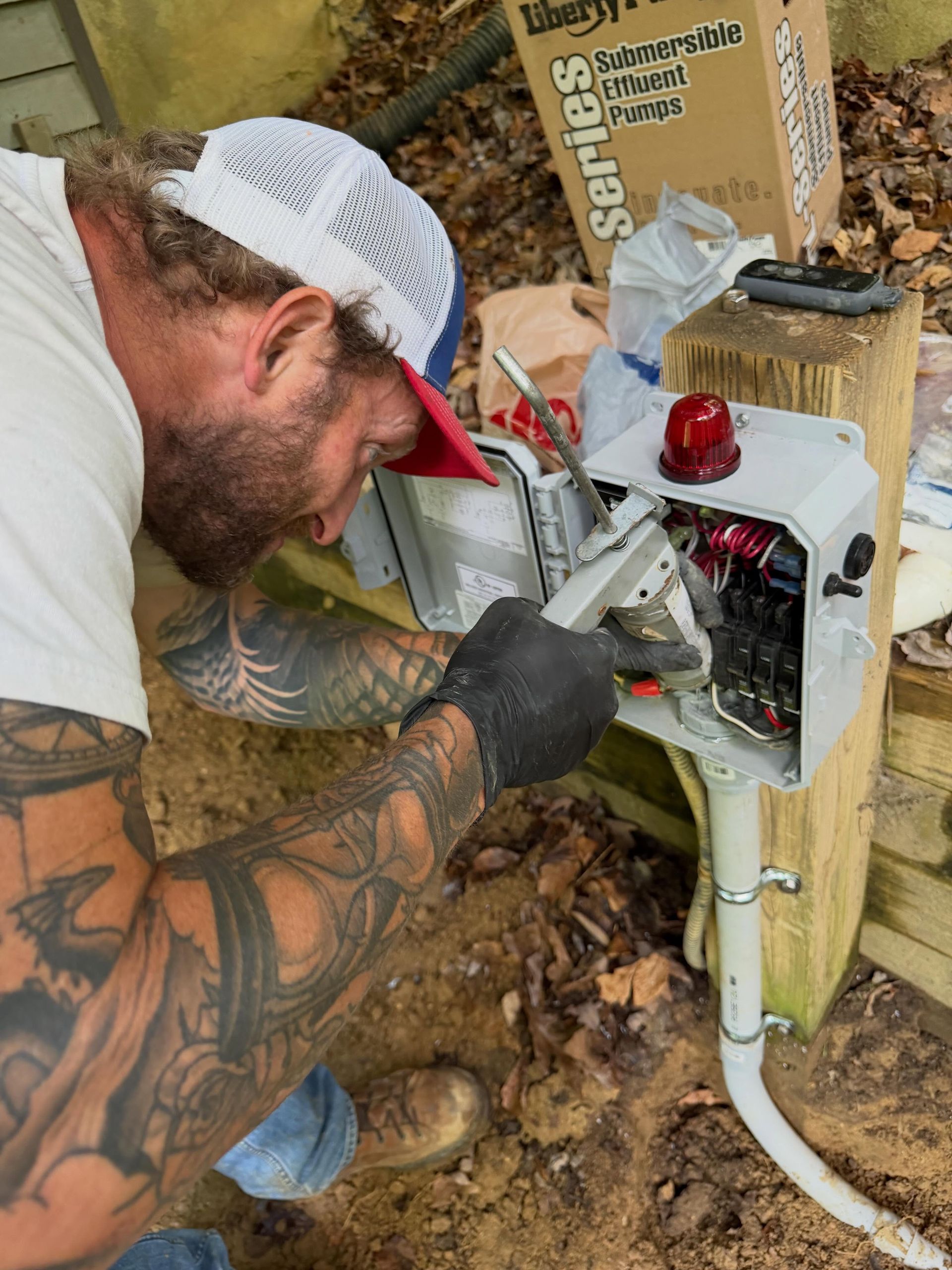 Worker repairing an open electrical box on a wooden post outdoors, with wires exposed and tools nearby