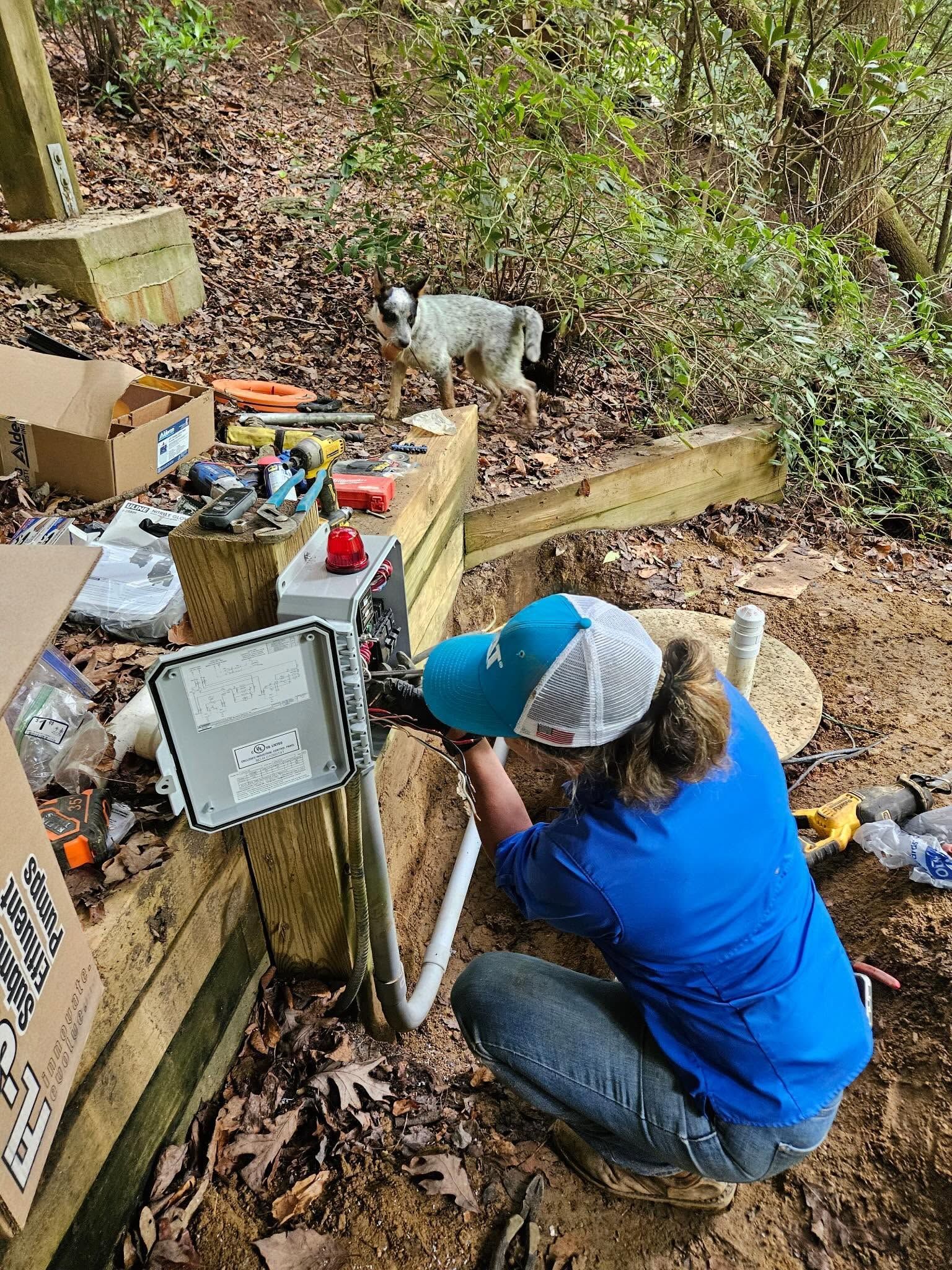 Person crouches by a broken appliance outdoors while a white dog stands nearby on a wooded hillside.
