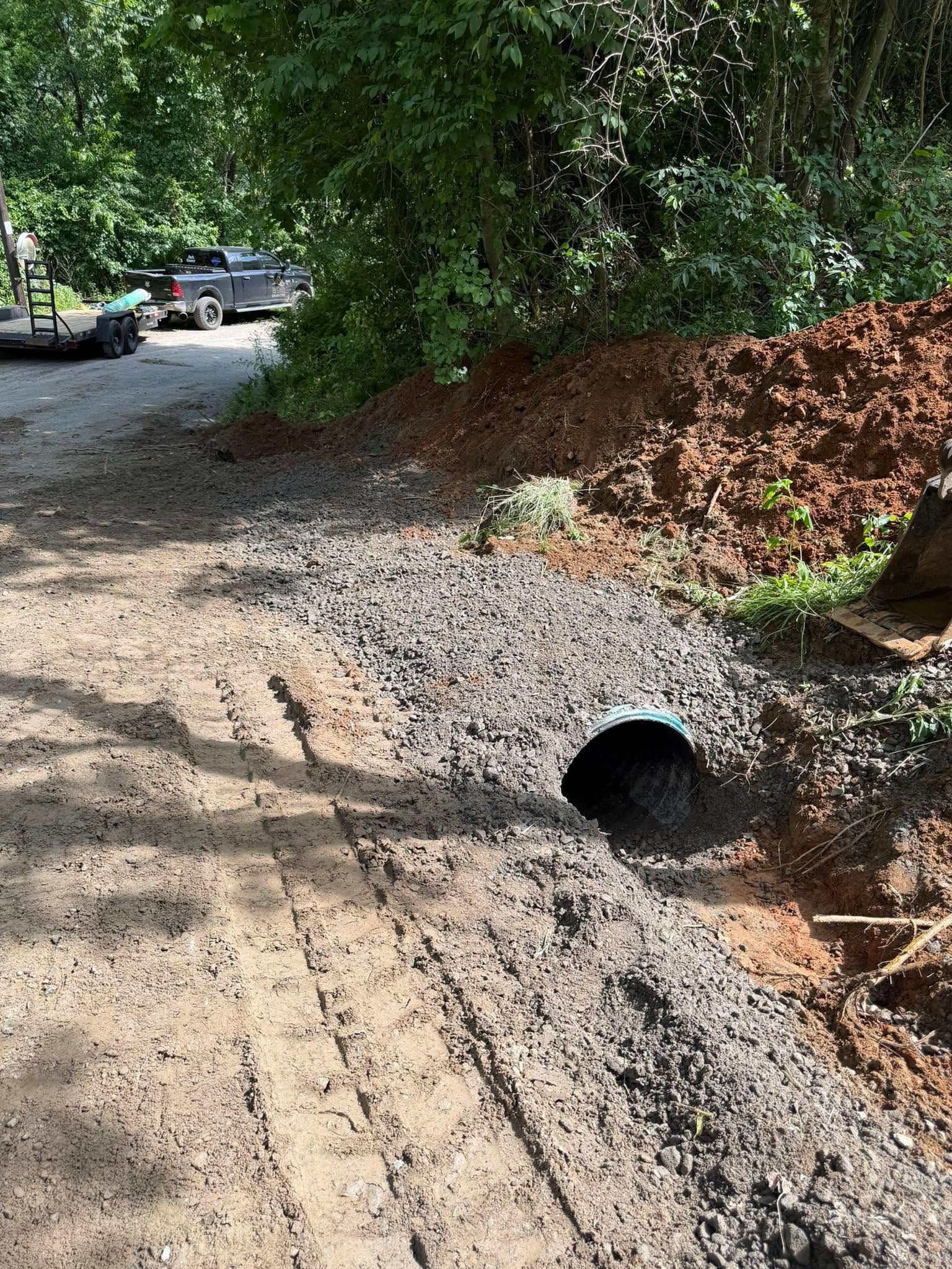 Gravel driveway with a drainage pipe beside a dirt embankment and trees; cars parked in the background.