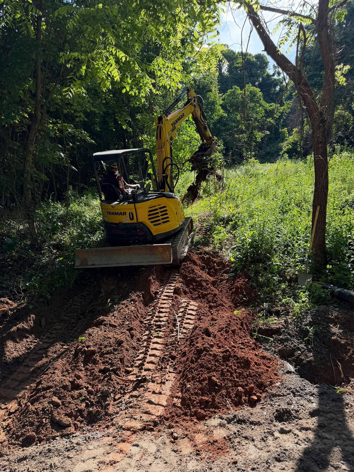 Yellow excavator clearing a muddy dirt path through dense green vegetation
