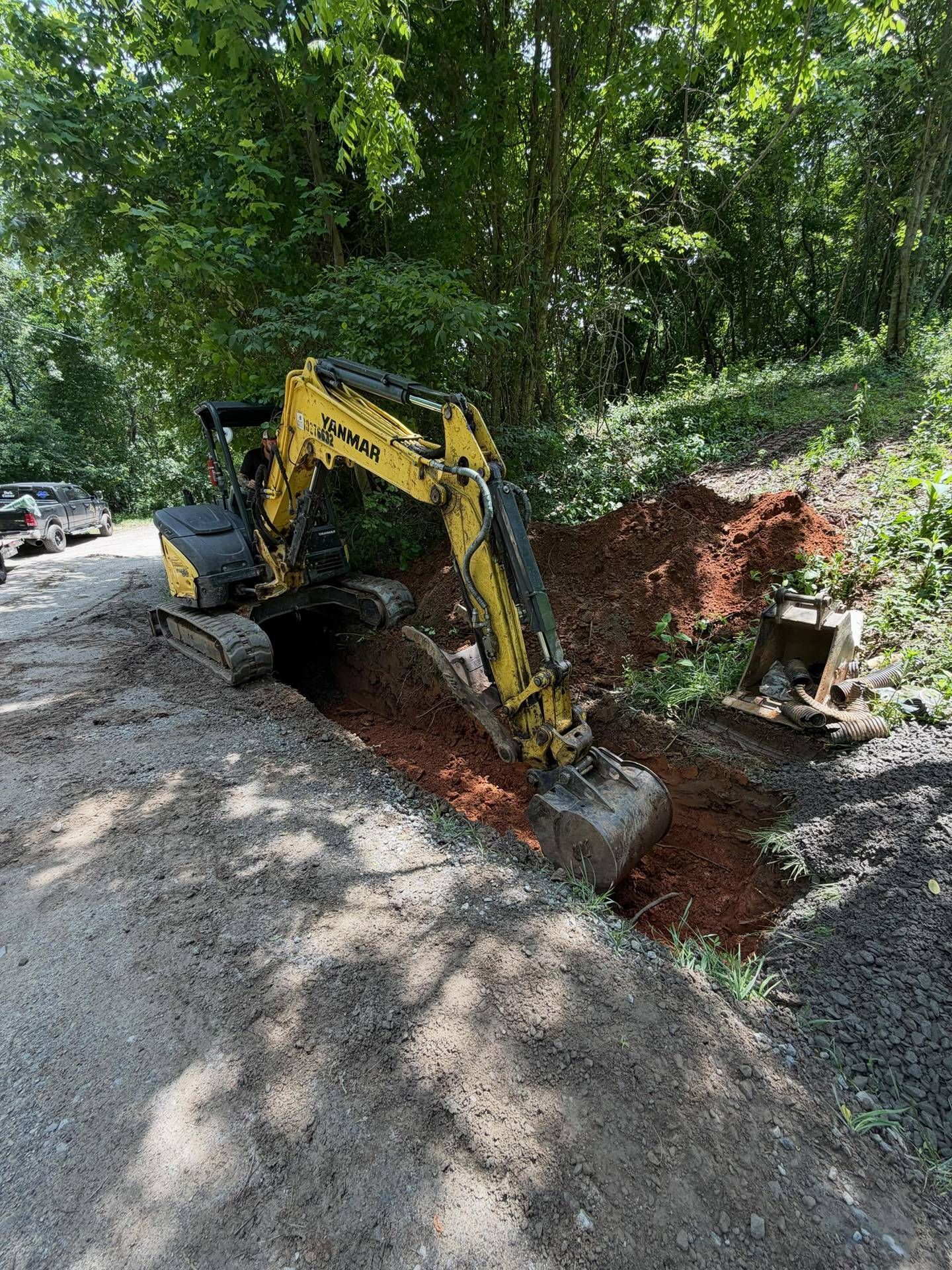 Yellow excavator digging a trench beside a wooded gravel road, with exposed red soil and a parked car nearby