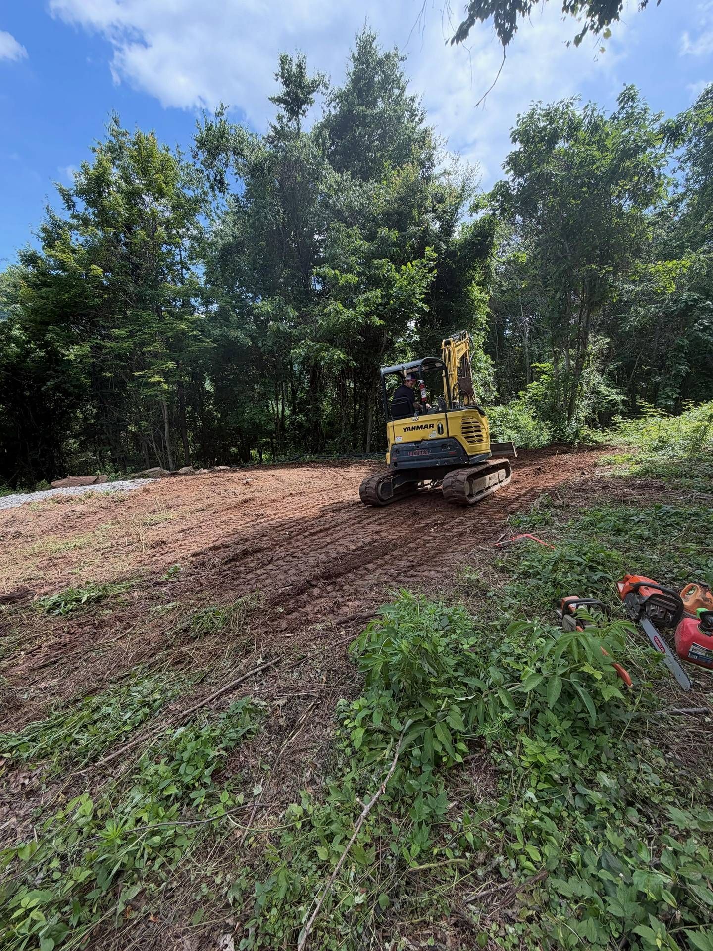 Yellow excavator clearing a dirt slope in a wooded area under a blue sky