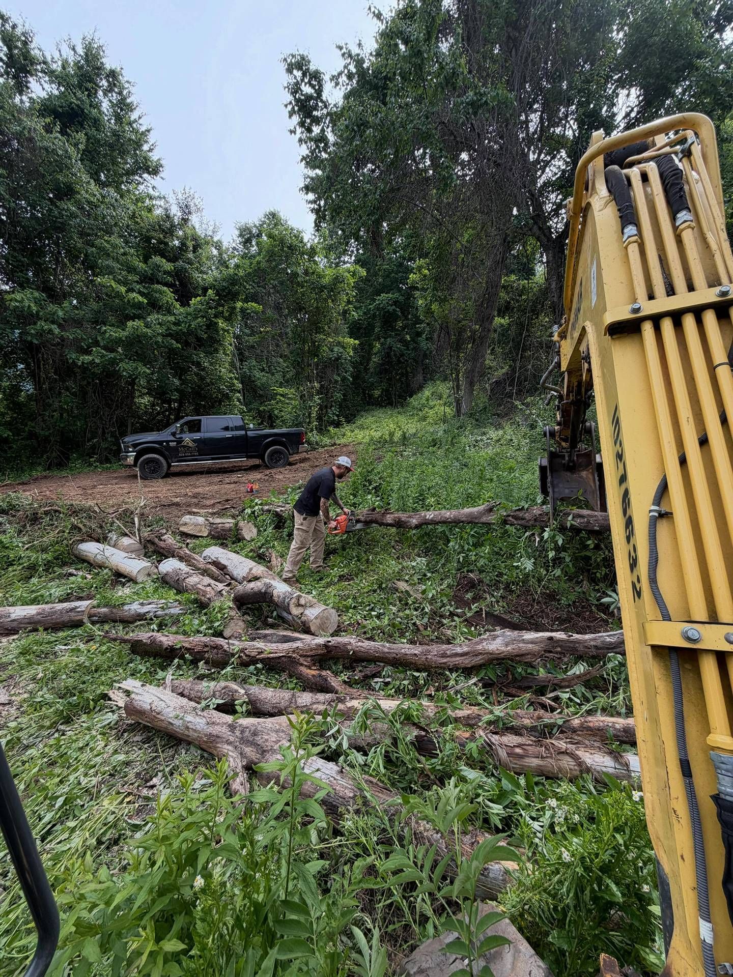 Cleared logs and a yellow excavator in a muddy, wooded clearing with a black pickup truck in the background