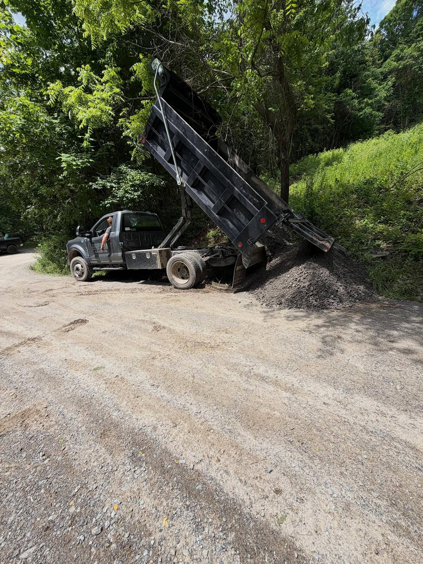 Dump truck unloading gravel on a dirt road beside trees and bushes
