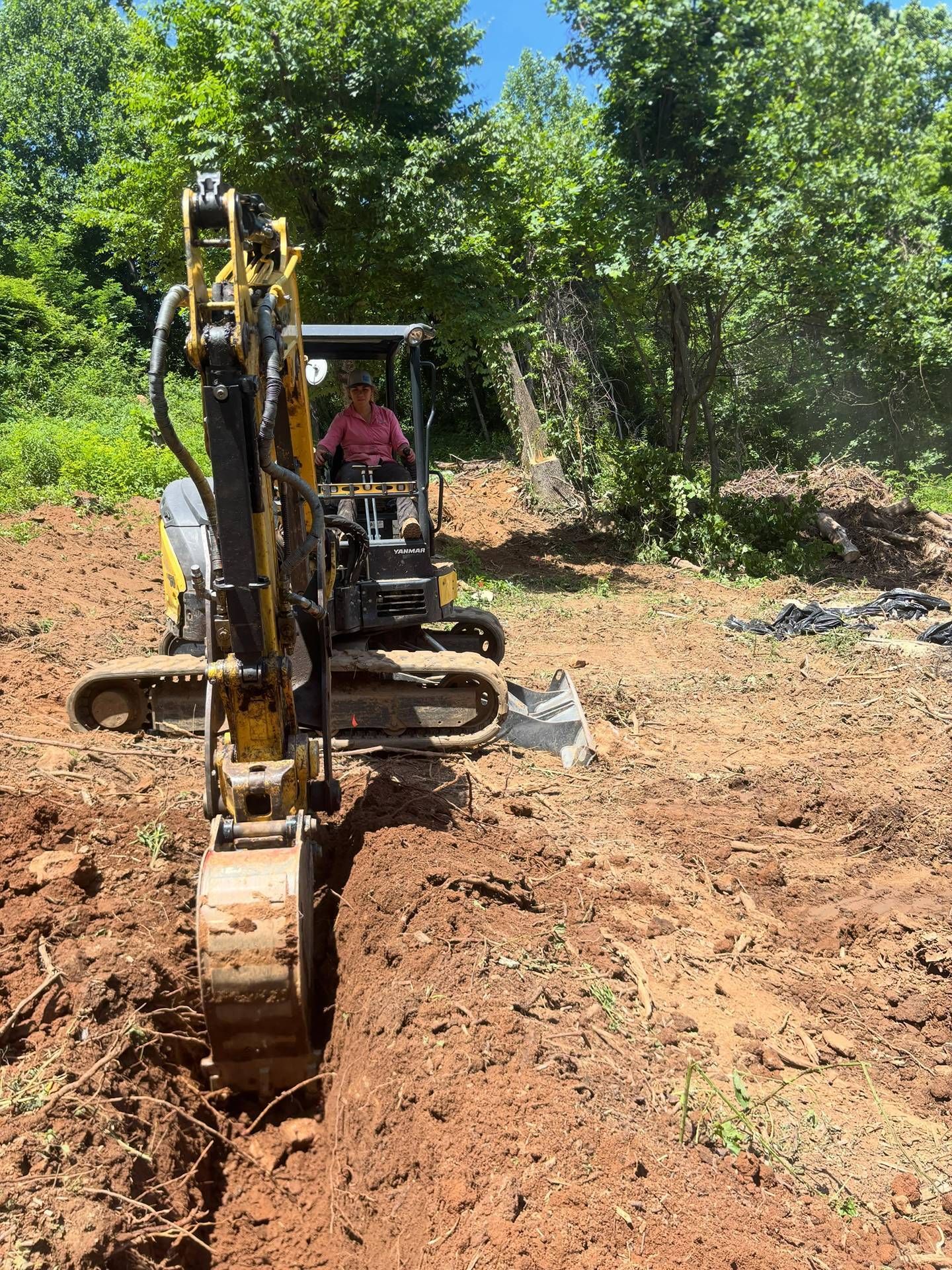 Small excavator digging a trench in a dirt clearing with trees in the background