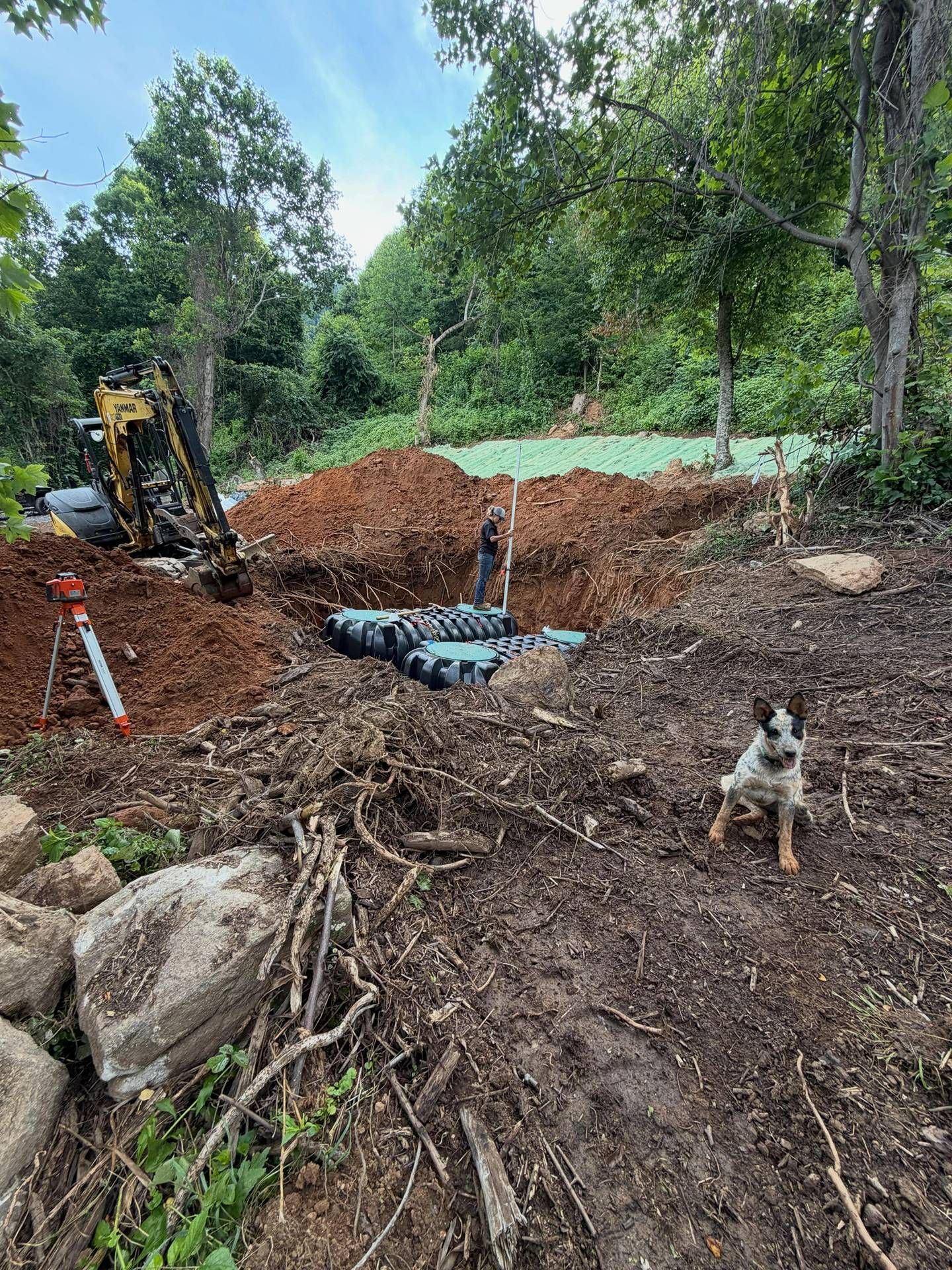 Excavator at a muddy construction site with exposed pipes, debris, and trees in the background
