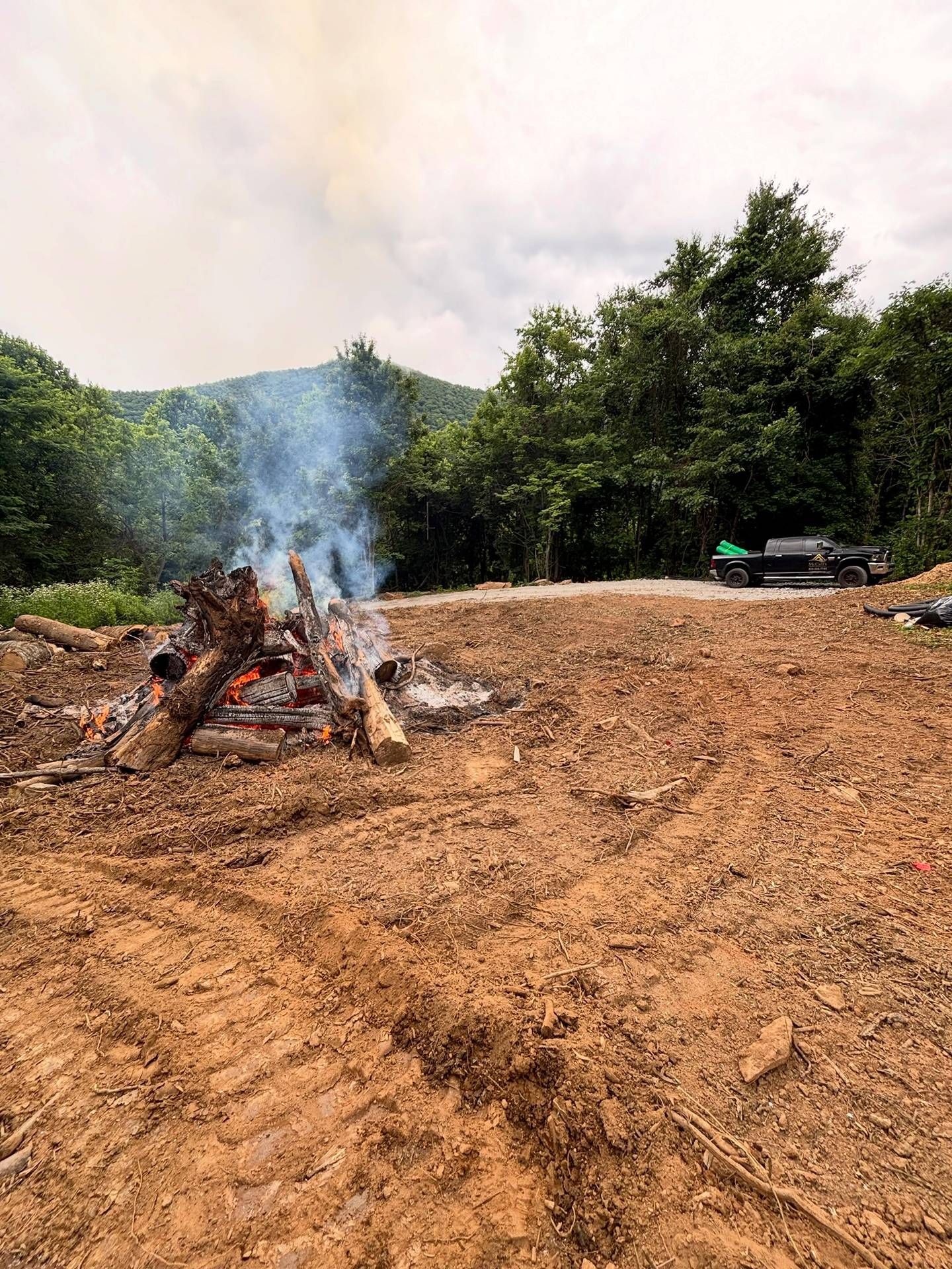 Smoke rises from a campfire on a muddy dirt clearing, with green trees and hills in the background.