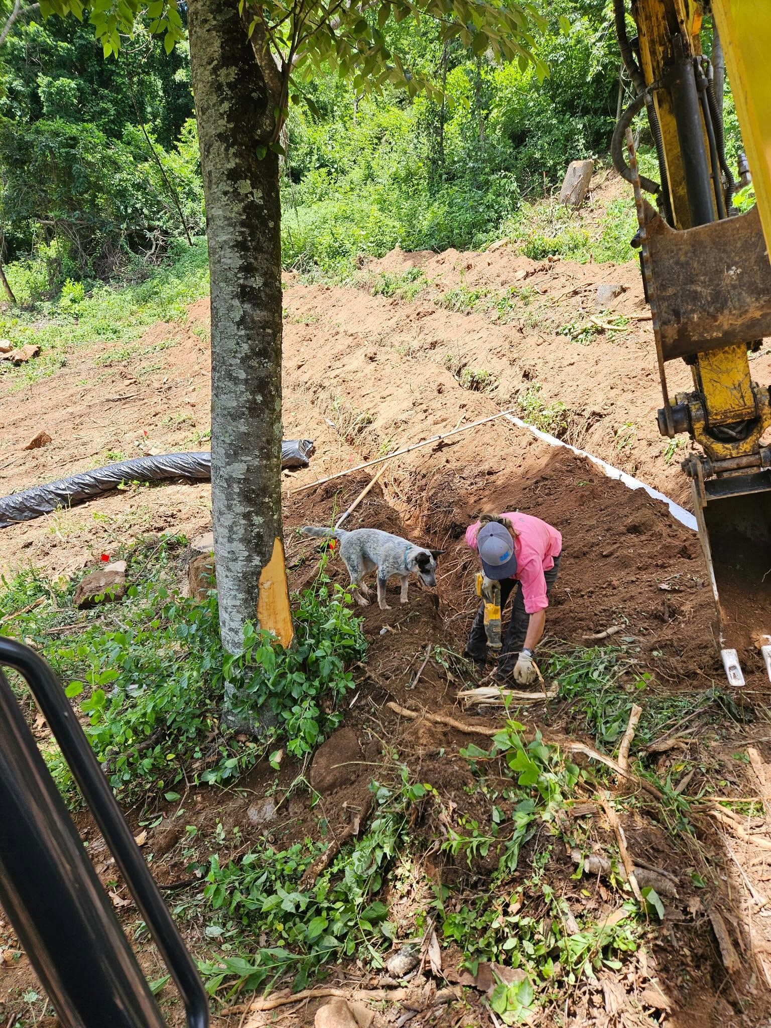 Worker in pink shirt digging in a muddy outdoor trench beside an excavator among trees