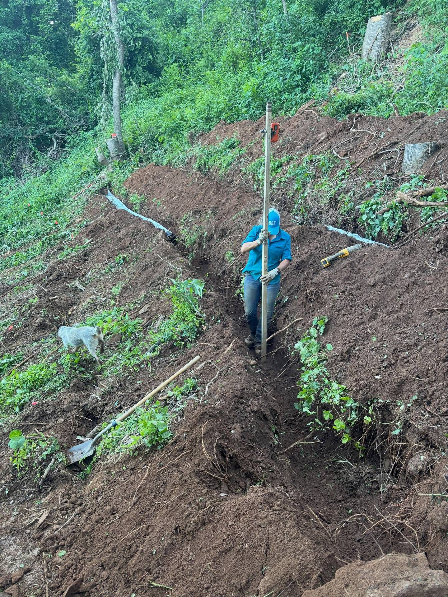 Person in a blue jacket standing in a muddy landslide area with exposed pipes on a forested slope