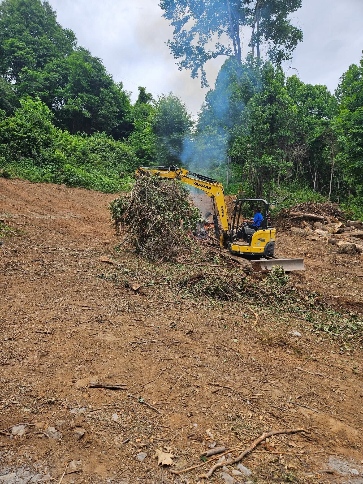 Yellow excavator clearing a dirt slope in a wooded area, with smoke rising in the background