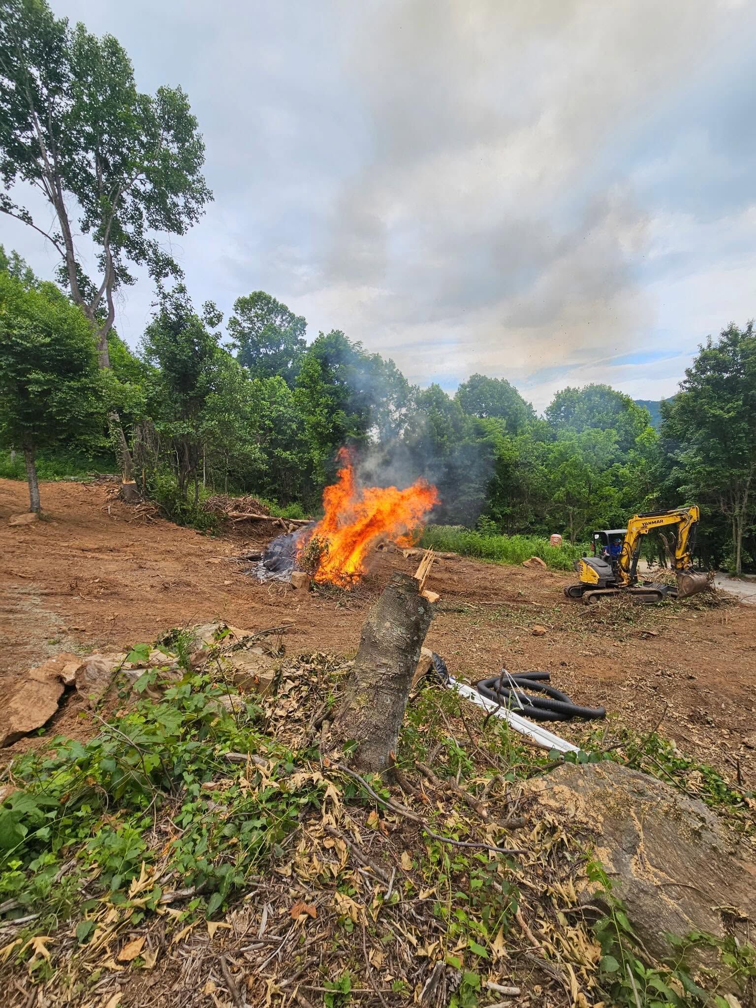 Small brush fire burning in a wooded clearing, with smoke and an excavator nearby