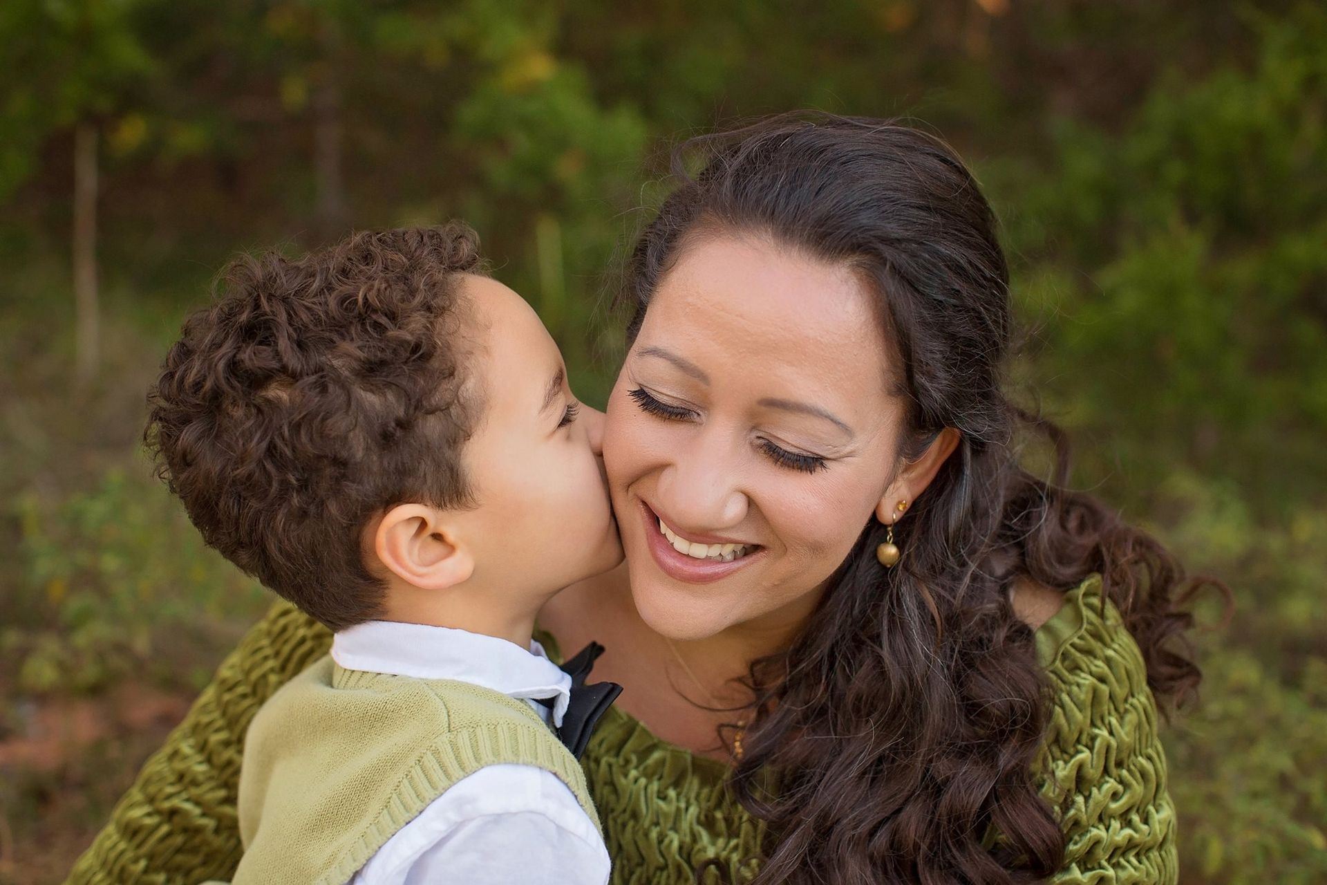 Son kissing mother's cheek; both smiling outdoors.
